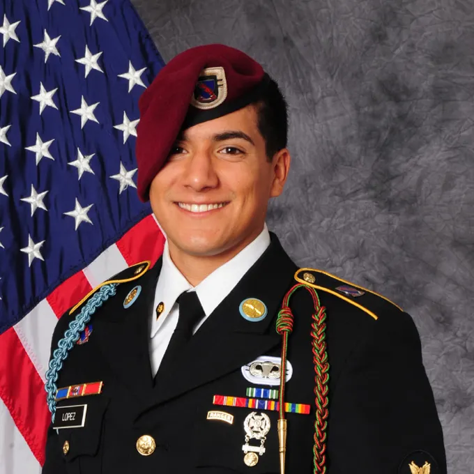 Mikey Lopez in uniform with medals, wearing a maroon hat. Behind him the flag of the United States.