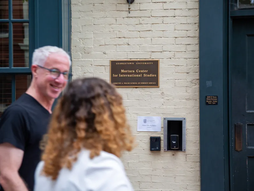 White brick building, focussed on the name plaque "Georgetown University Mortara Center for International Studies School of Foreign Service" Our of focus a man and a woman walking in front of the building.