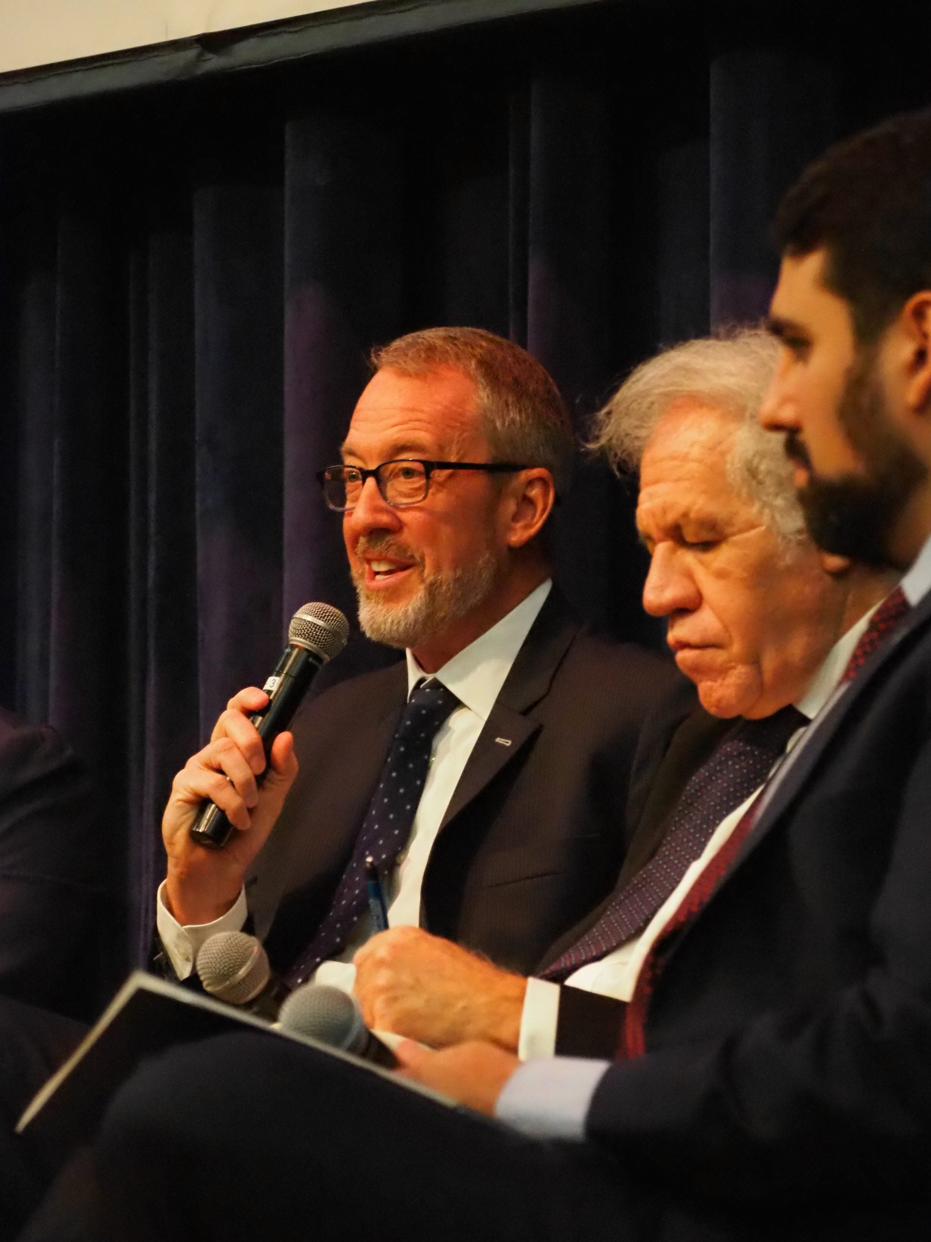 Photo of three panelists sitting. U.S. Ambassador James Story, at the left is answering questions with a microphone.