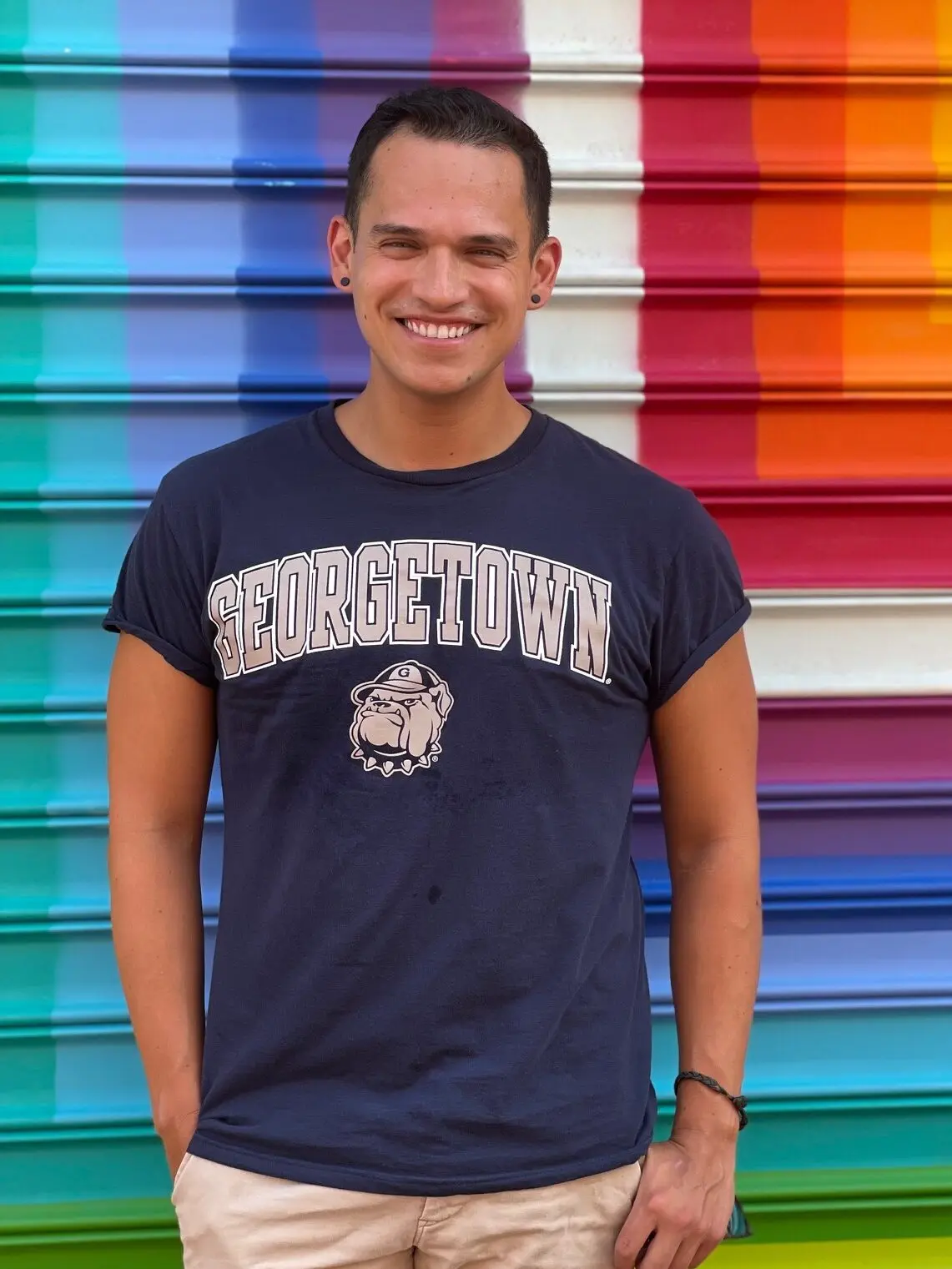 Brian Castro, wearing a Georgetown t-shirt and smiling in front of a rainbow colored wall.