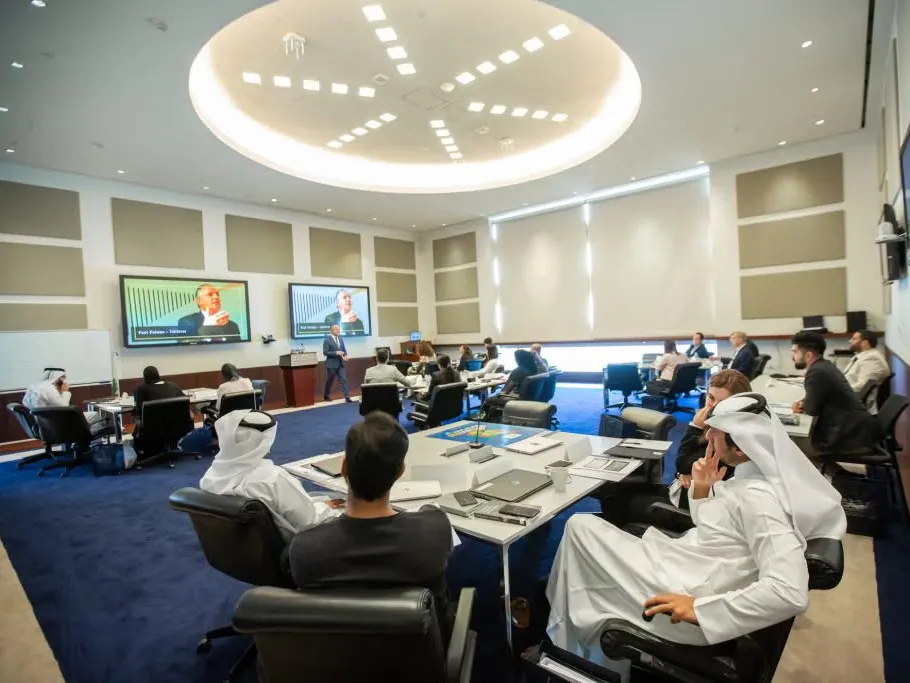 Classroom in Qatar, men and women sitting around tables, taking notes, looking at a virtual speaker on screens at the front of the classroom.