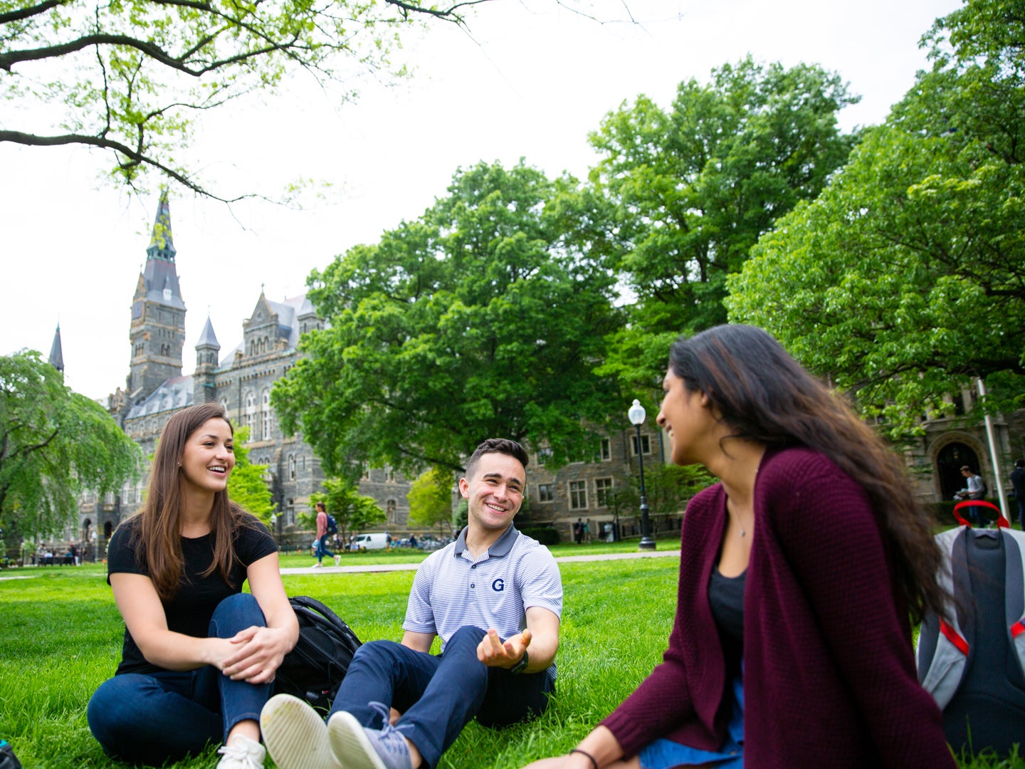 Three students sitting on the green Georgetown University campus grass. They are speaking to each other, relaxed, smiling.