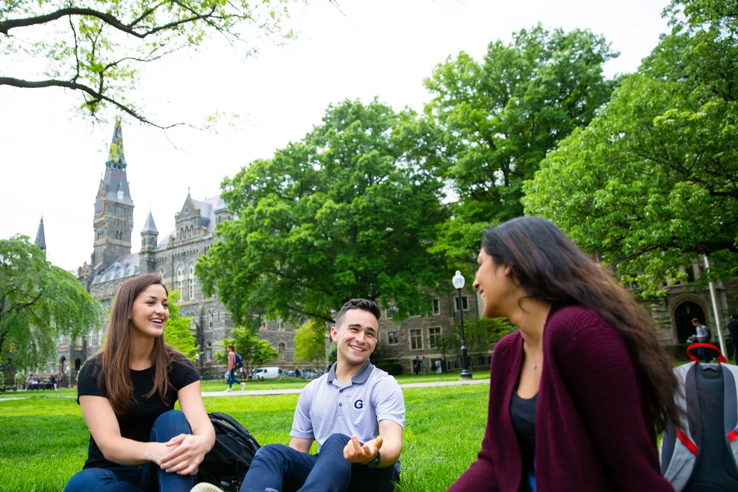 Three students sitting on the green Georgetown University campus grass. They are speaking to each other, relaxed, smiling.