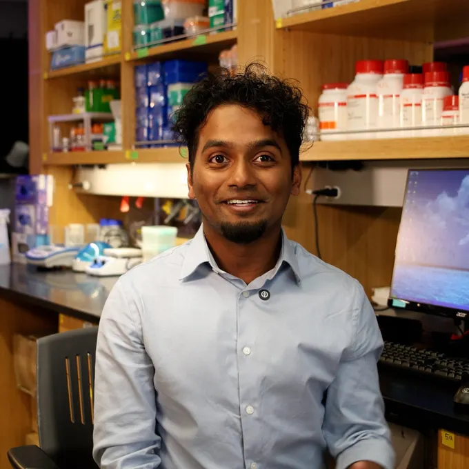 Zachariah John smiling, wearing a light blue buttoned up shirt. He is sitting in a lab, behind him shelves of equipment and materials.