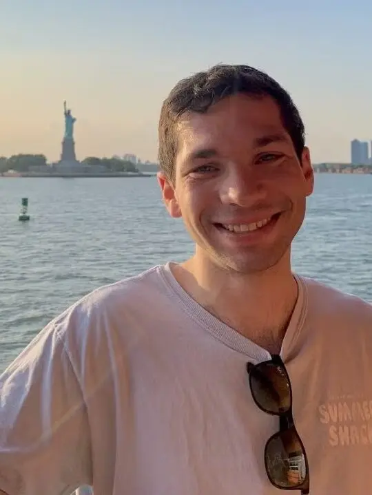 Caleb Diamond standing with Statue of Liberty in background, wearing a light gray t-shirt.