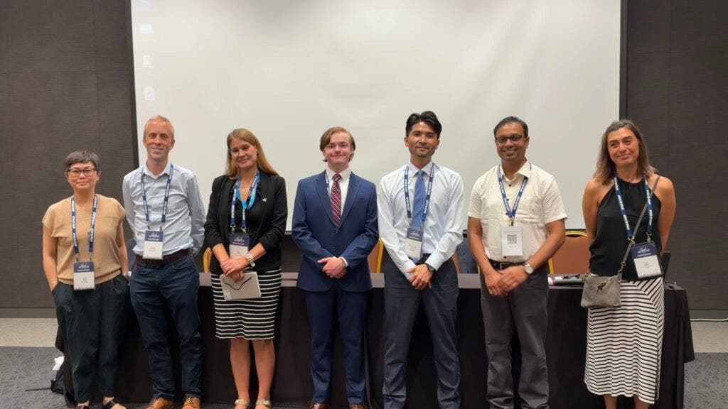 Harrison Brown stnads with a group of people in front of a white screen in a conference room.