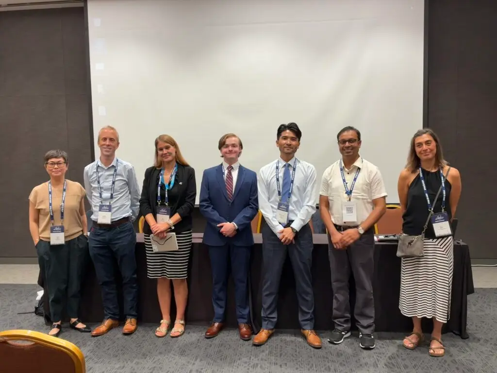 Harrison Brown stnads with a group of people in front of a white screen in a conference room.