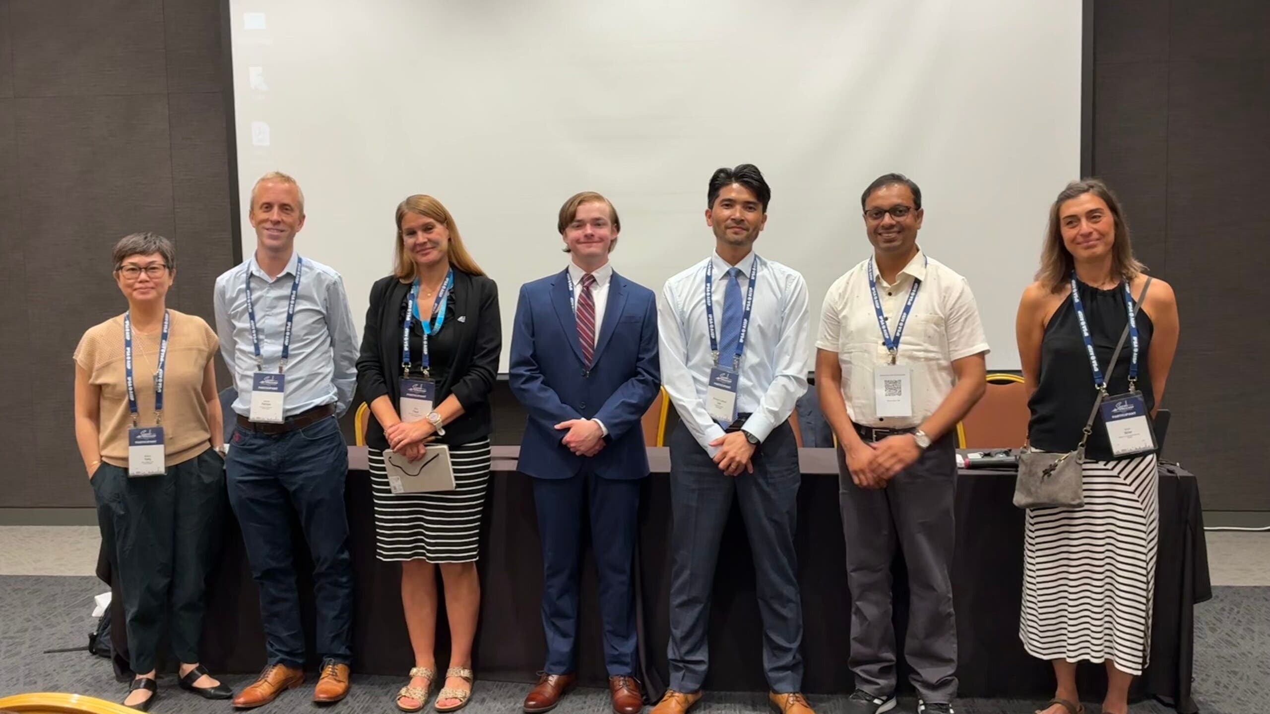 Harrison Brown stnads with a group of people in front of a white screen in a conference room.