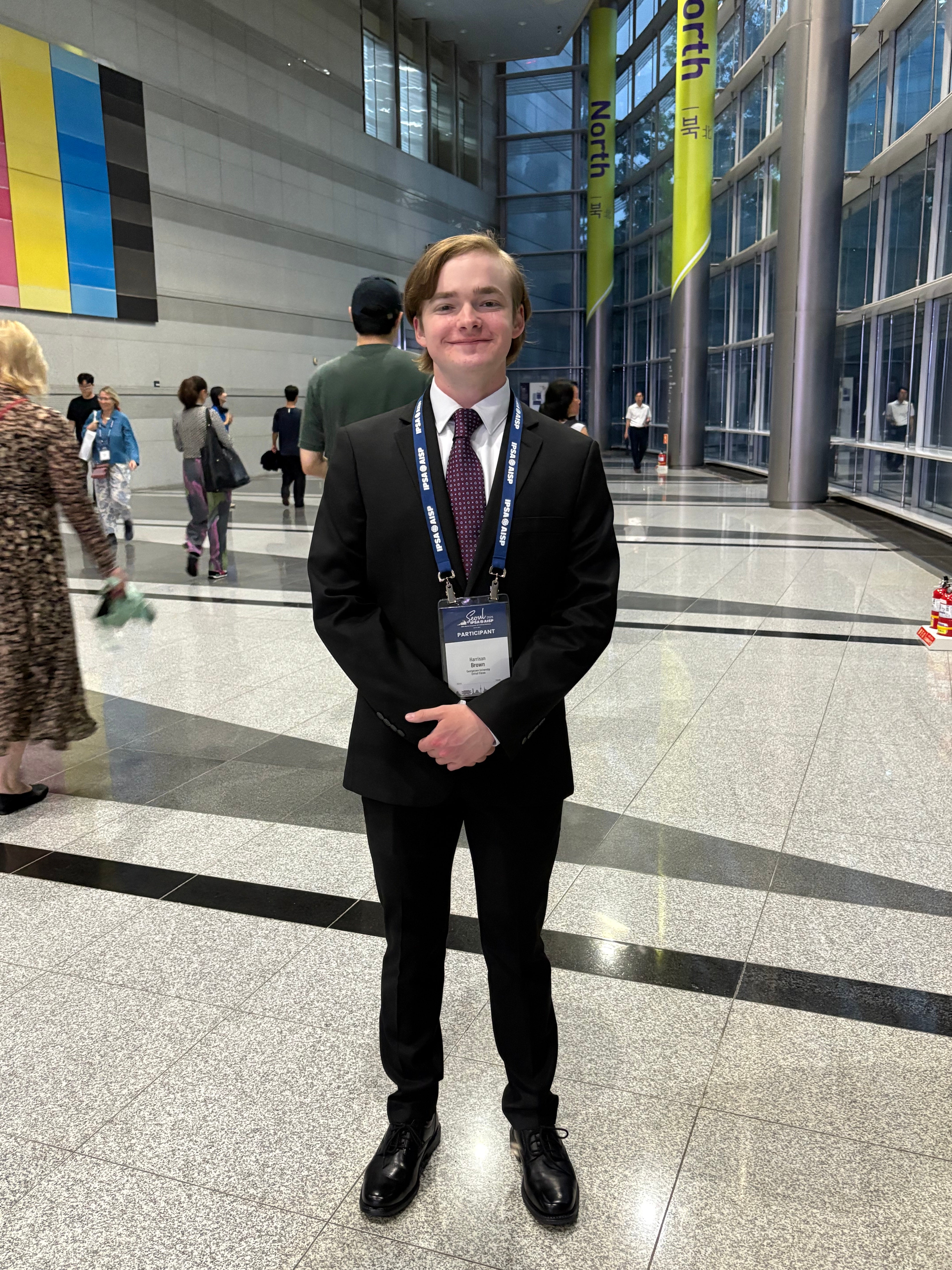 Harrison Brown stands in a suit wearing a lanyard in a wide open space with people in the background.