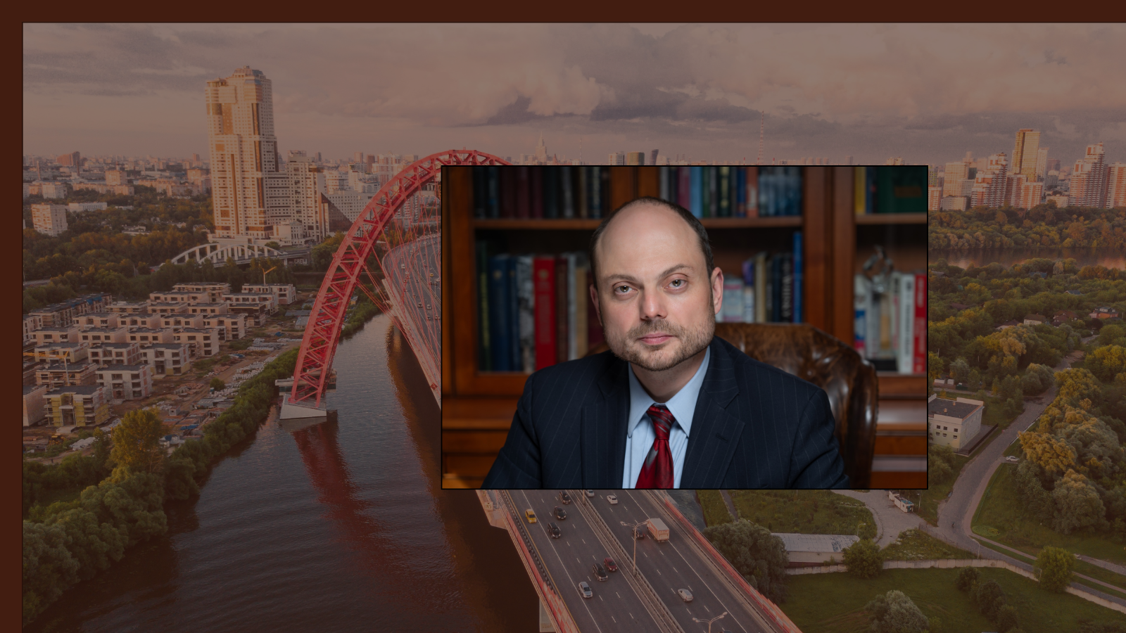 A balding man in a blue suit with a red tie, seated in front of bookshelves with glass doors