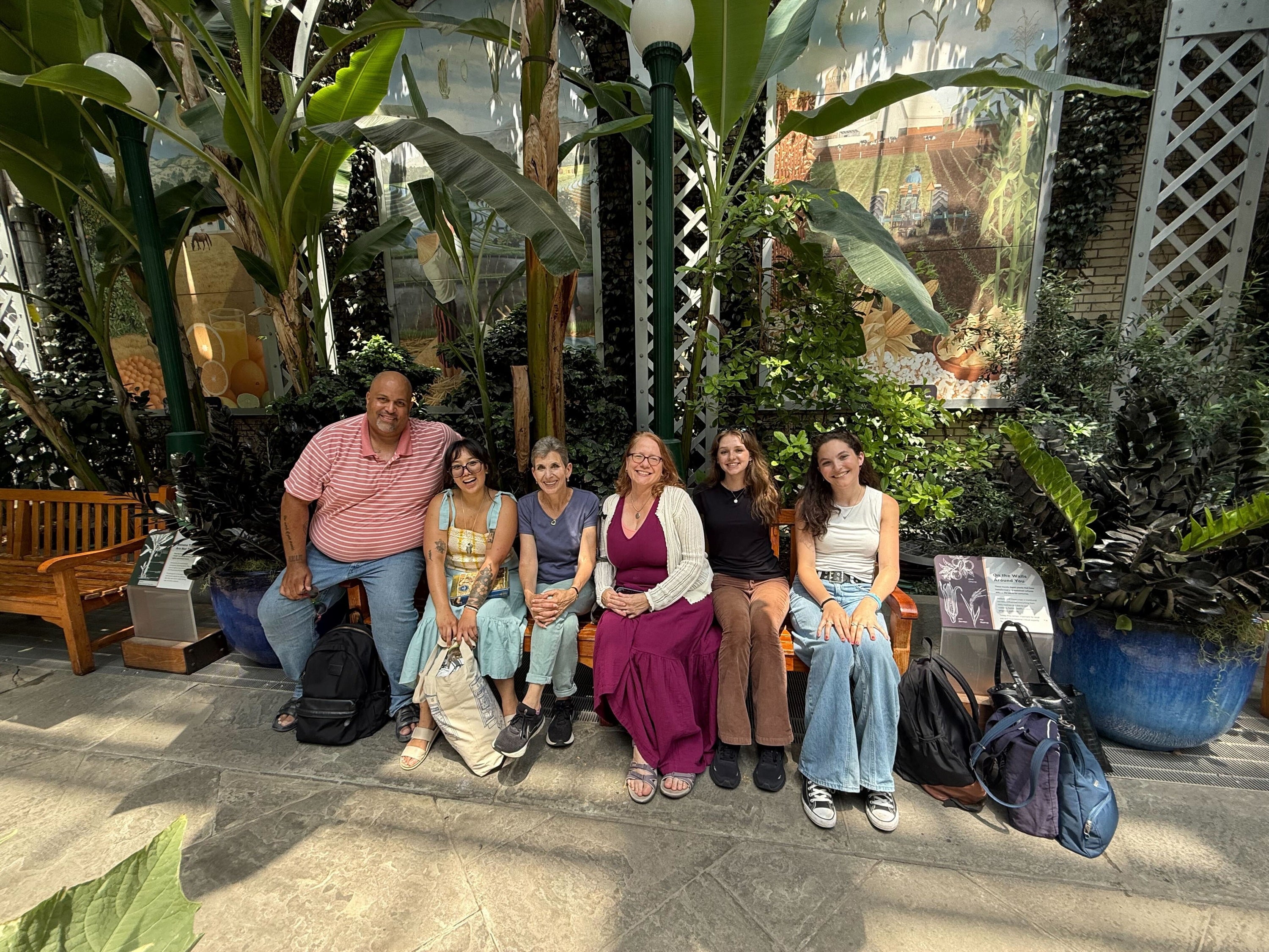 Six individuals seated on a bench in a lush indoor garden setting, smiling at the camera.