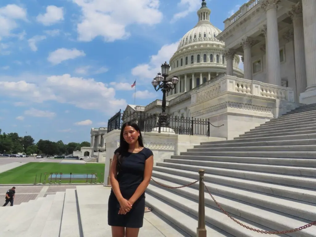 A woman with dark hair wearing a black dress standing in front of the United States Capitol Building on a sunny day.