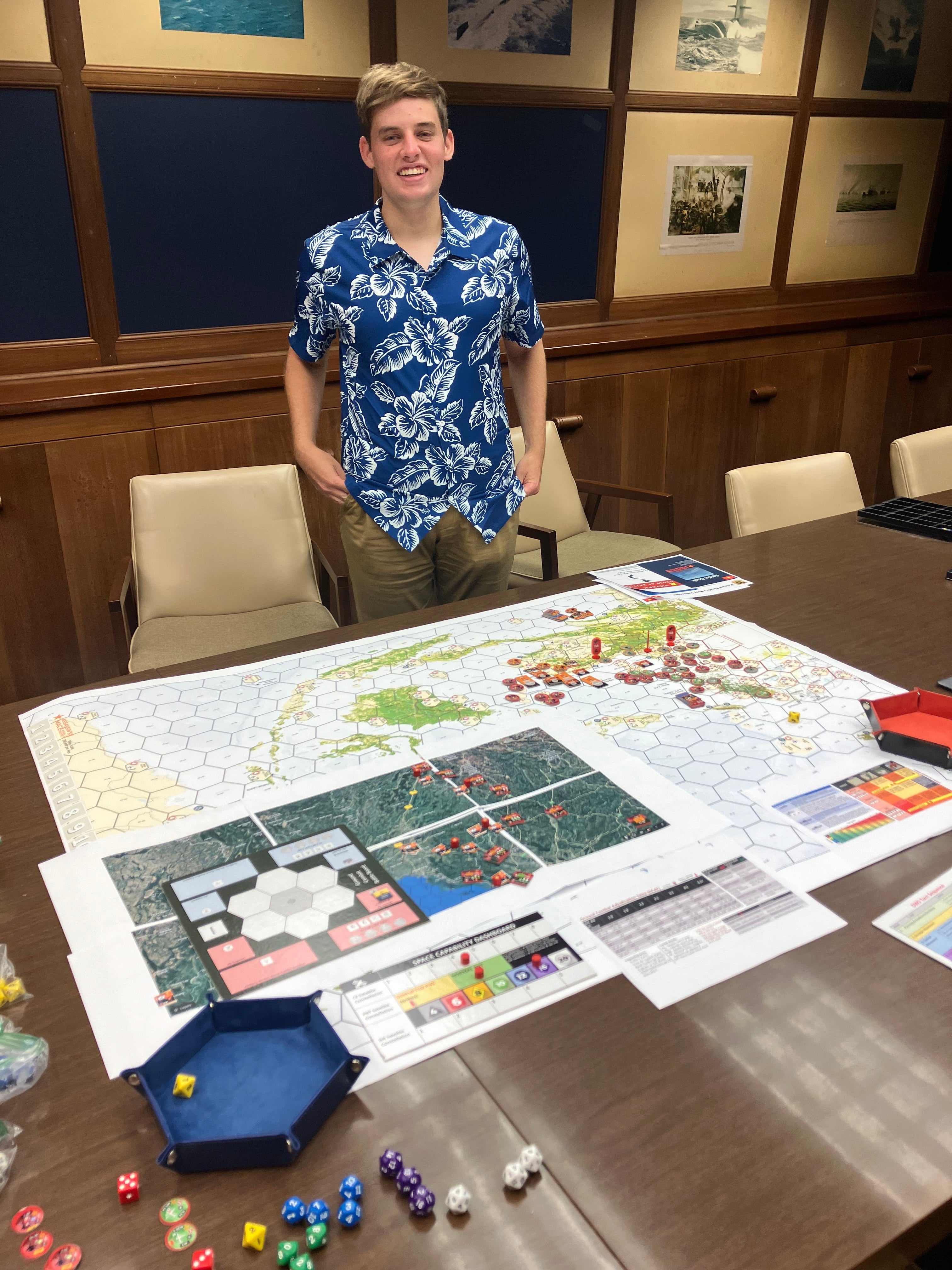 Person standing behind a table with a complex board game setup, featuring maps, colorful dice, and game tokens.