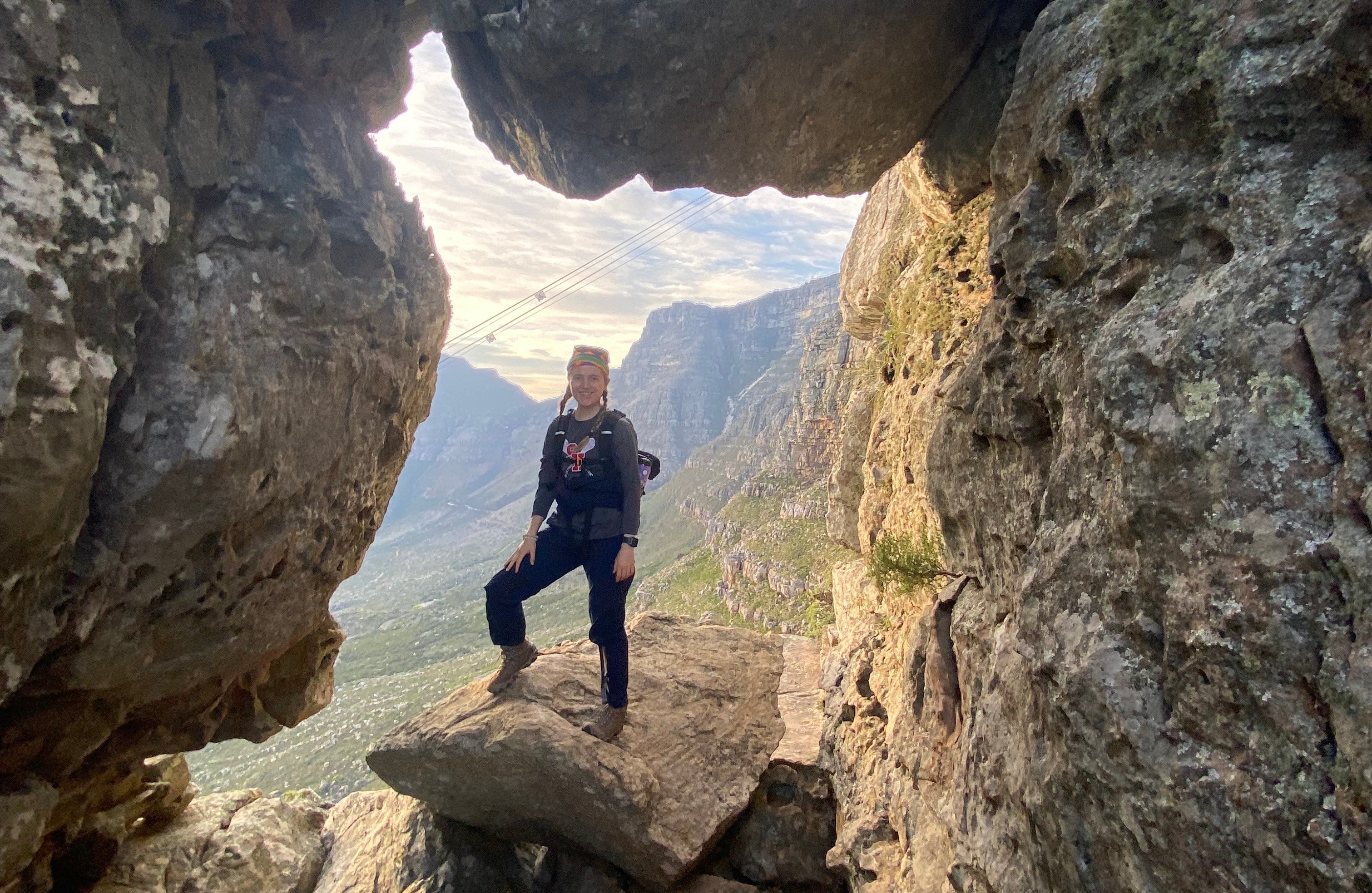 Person standing in a rocky cave opening with a view of Table Mountain and cable cars in the background.