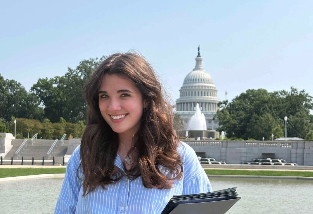 Woman with long brown hair wearing a blue shirt smiling with a folder in hand, standing in front of the United States Capitol building on a sunny day.