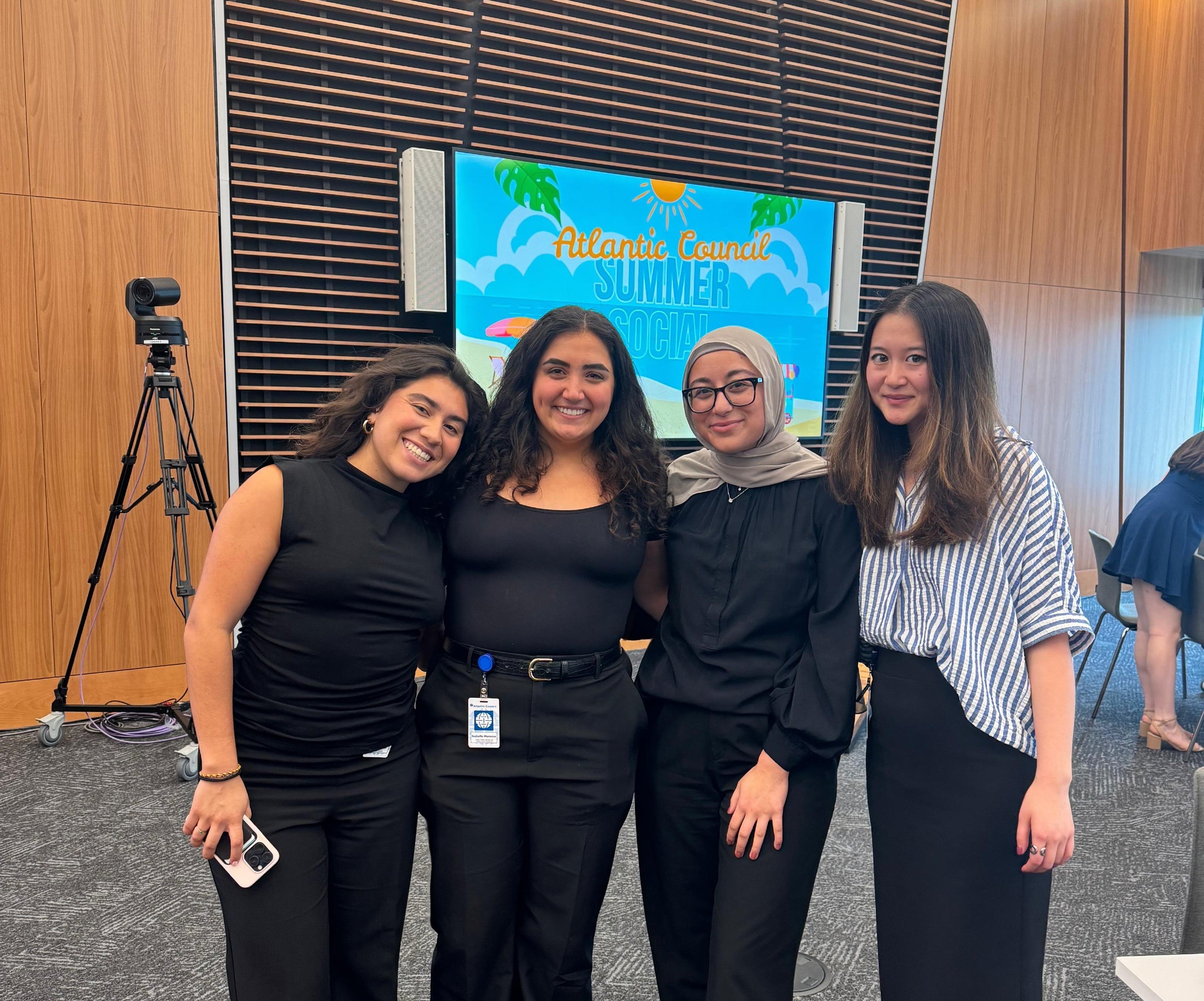 Three people smiling for a photo at an event. Two are dressed in black and one in blue, standing in front of a digital display and wooden paneling.