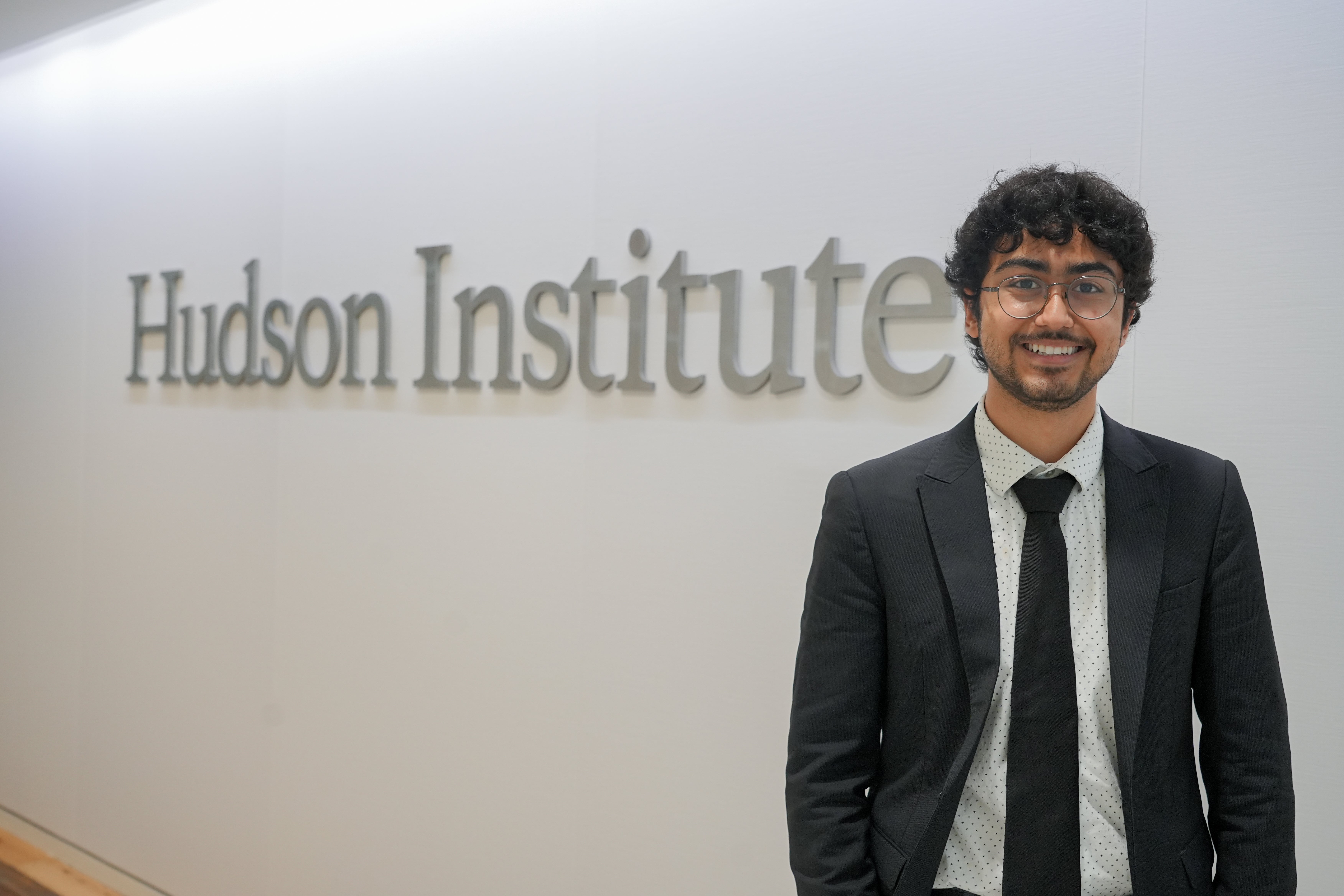 Person in a business suit smiling in front of a wall with the Hudson Institute logo.