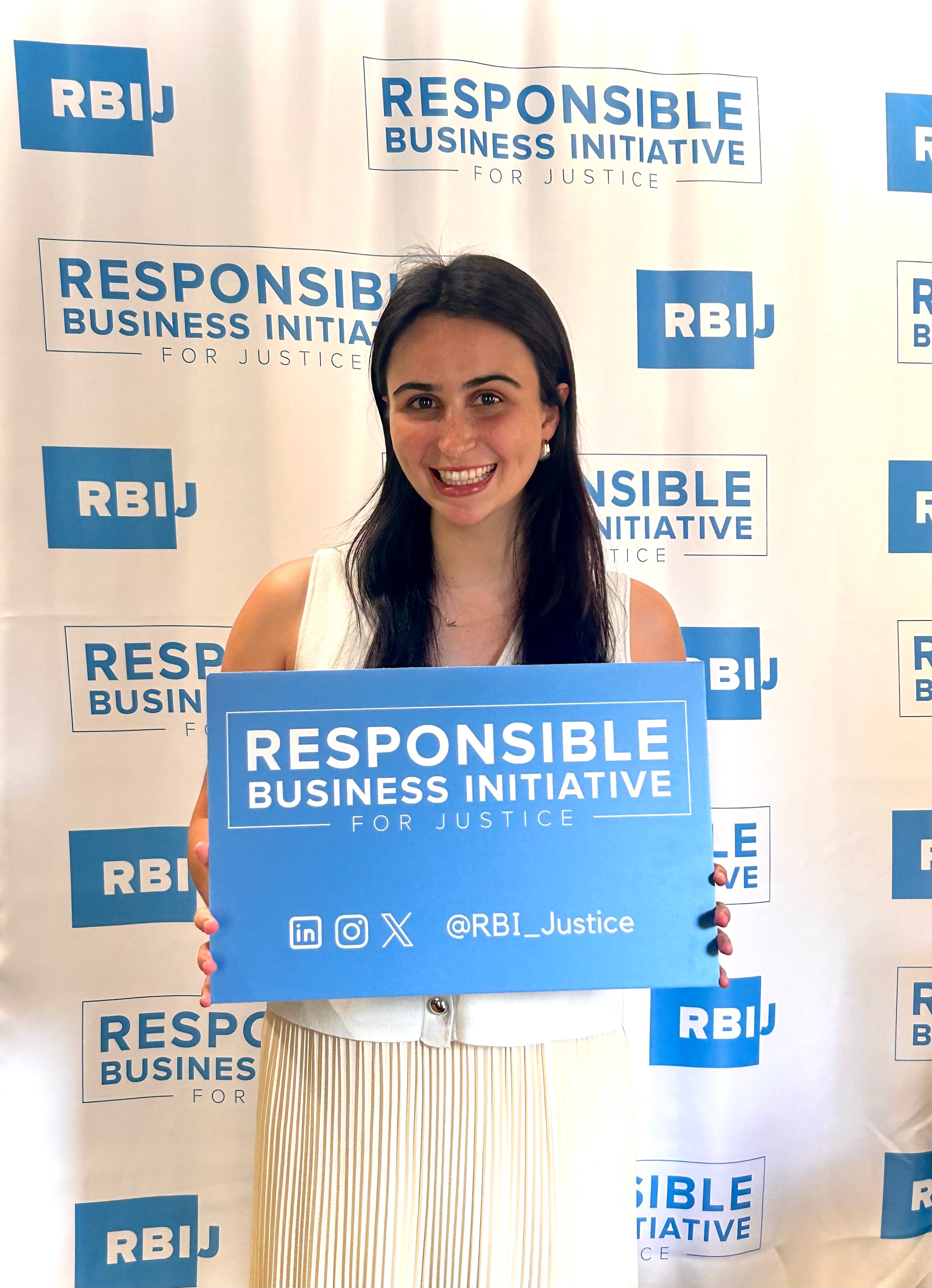 Individual holding a sign with the logo of the Responsible Business Initiative for Justice, standing in front of a banner with multiple RBI logos.