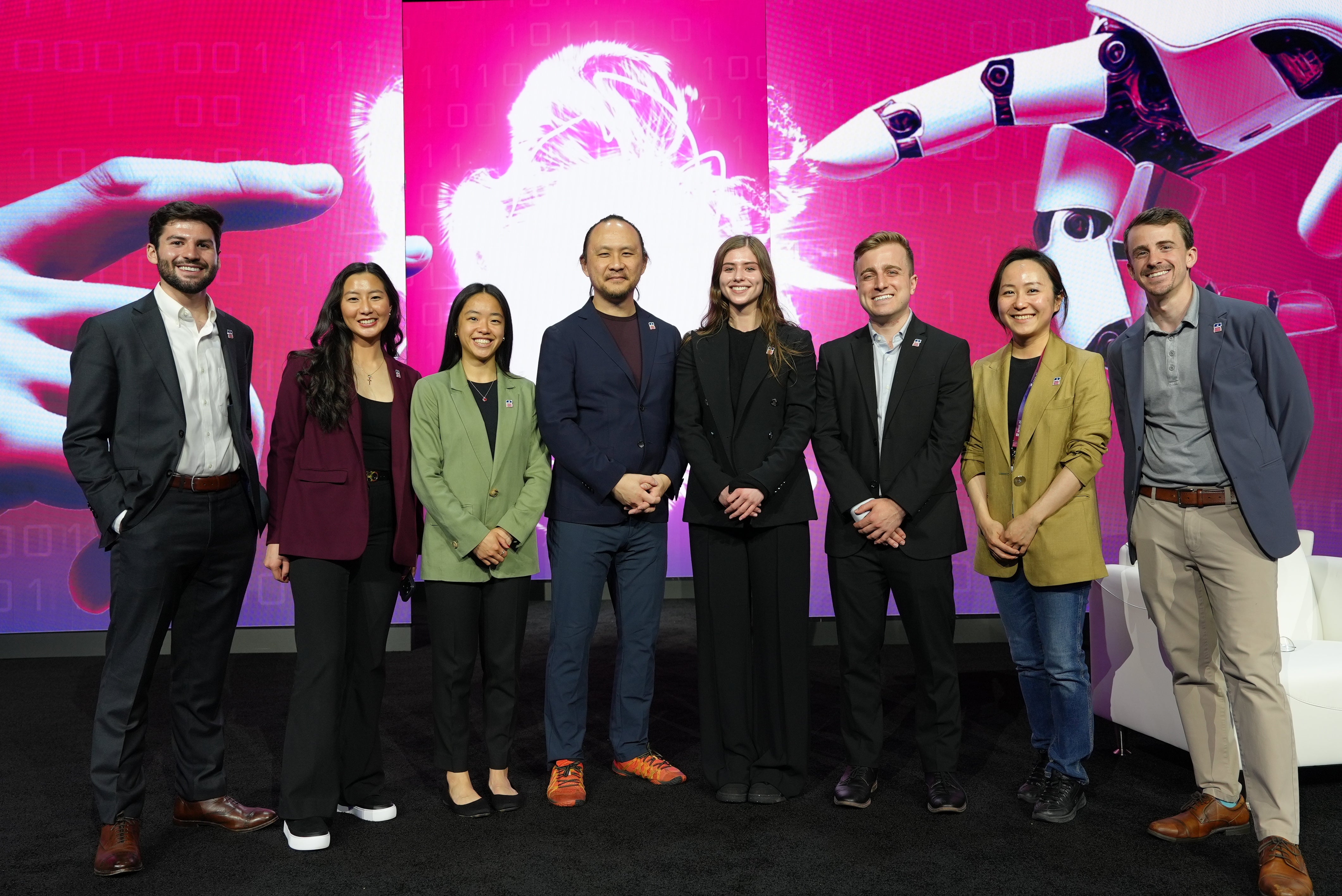 Group of eight professionals smiling and standing together at a tech event with a vibrant pink digital background featuring abstract graphics.
