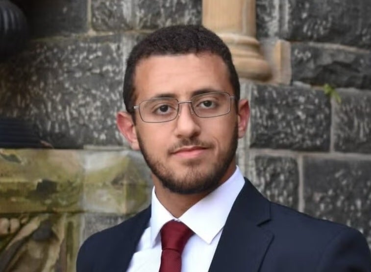 Portrait of a person wearing glasses, a suit, and a red tie, standing in front of a stone building.