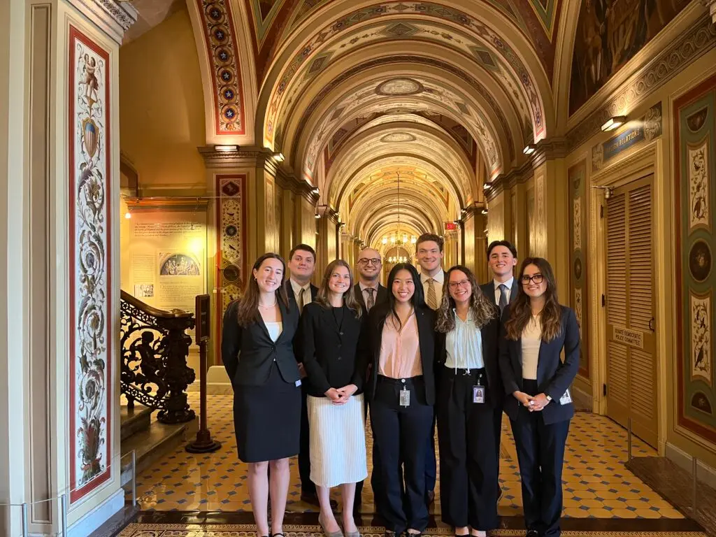 A group of professional individuals posing in a corridor of the United States Capitol building, featuring ornate architectural details and colorful patterns.