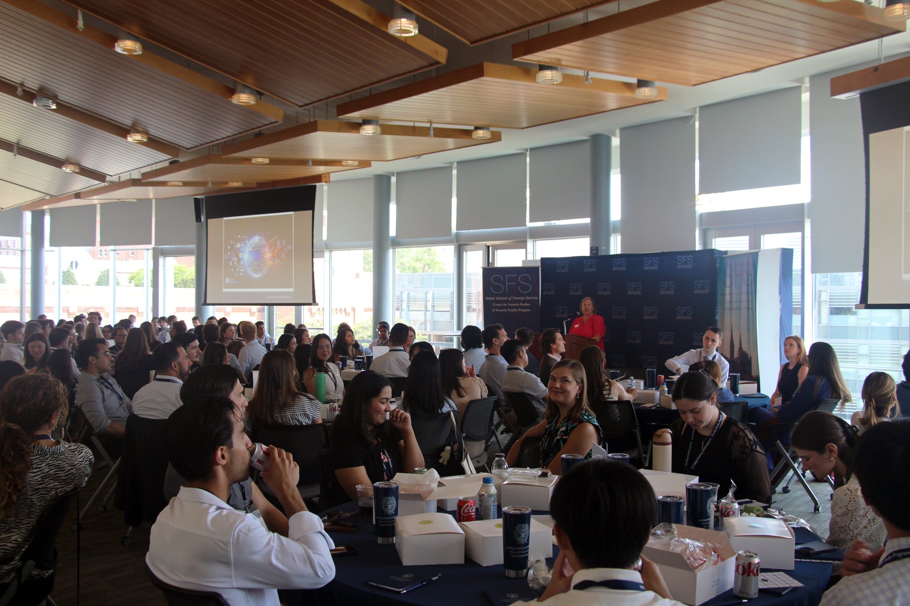 Students sitting in round tables at a bright event venue, while a person speaks on stage