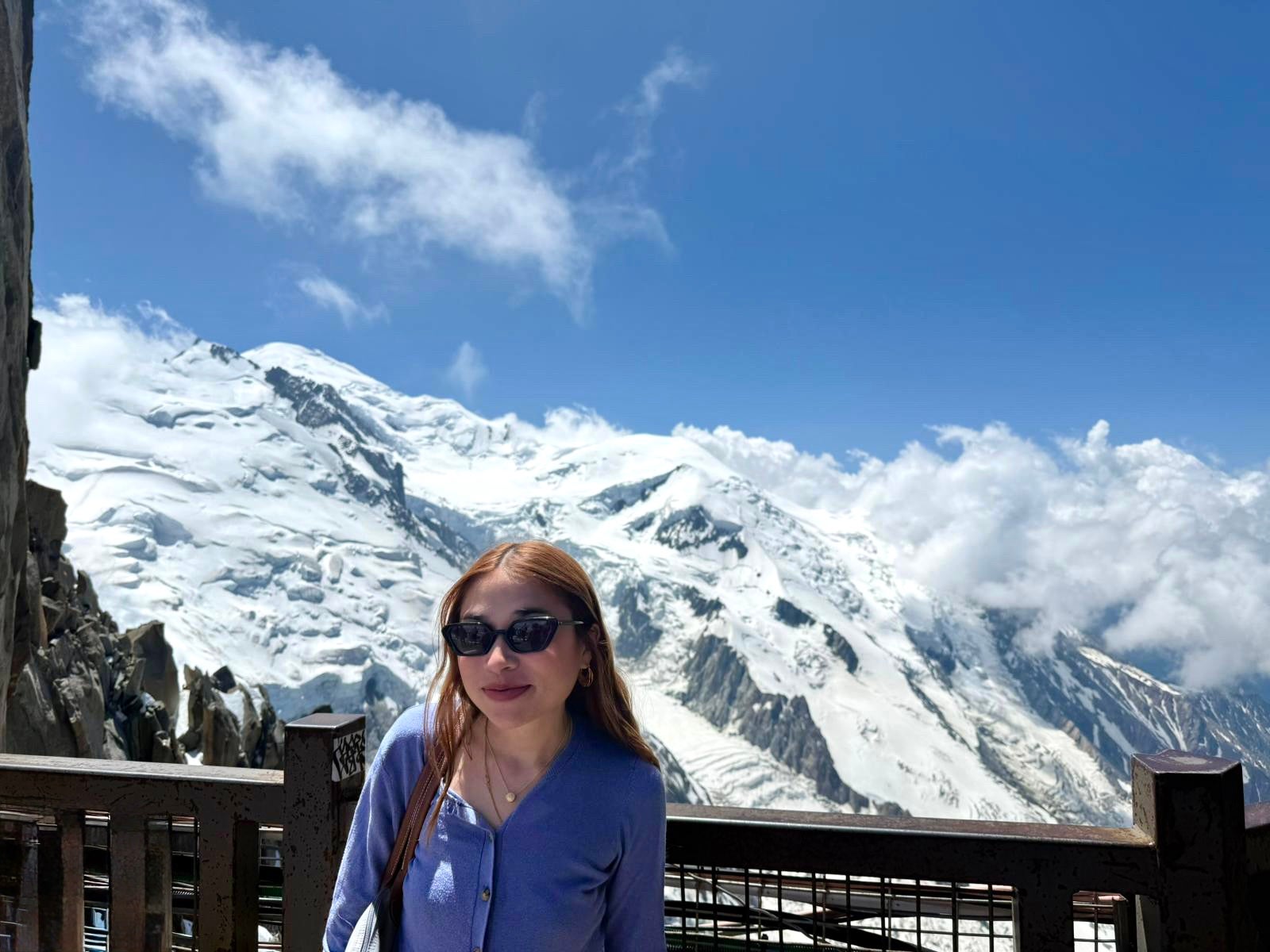 Woman wearing a purple sweater and black sunglasses standing on an observation deck with a view of Mont Blanc covered in snow and surrounded by clouds in the background.