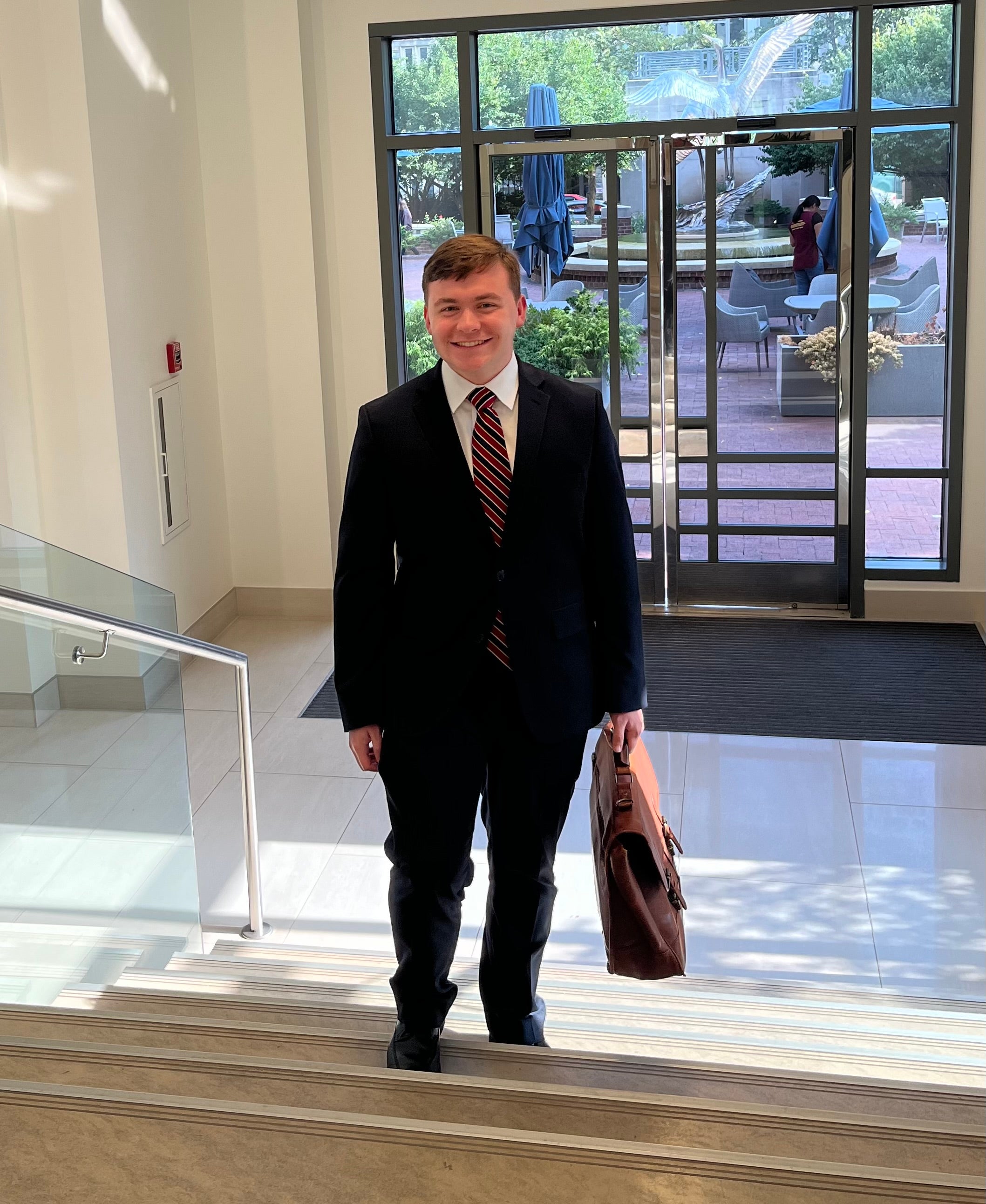 Person in a dark suit and striped tie smiling, holding a briefcase, standing indoors with glass doors and a sunny courtyard visible in the background.
