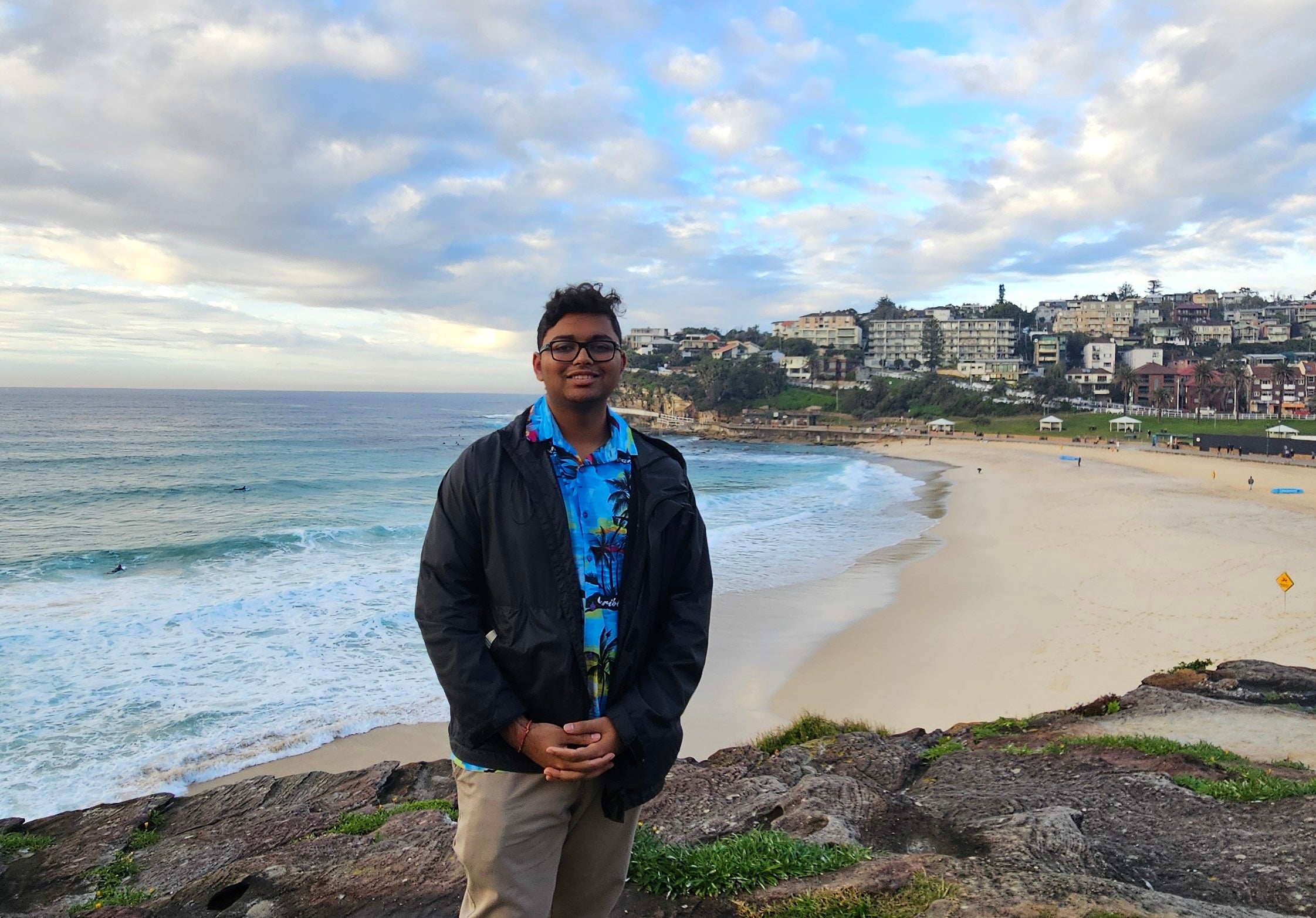 Person standing in front of the beach at Bondi Beach, with sand, ocean, and a cloudy sky in the background.