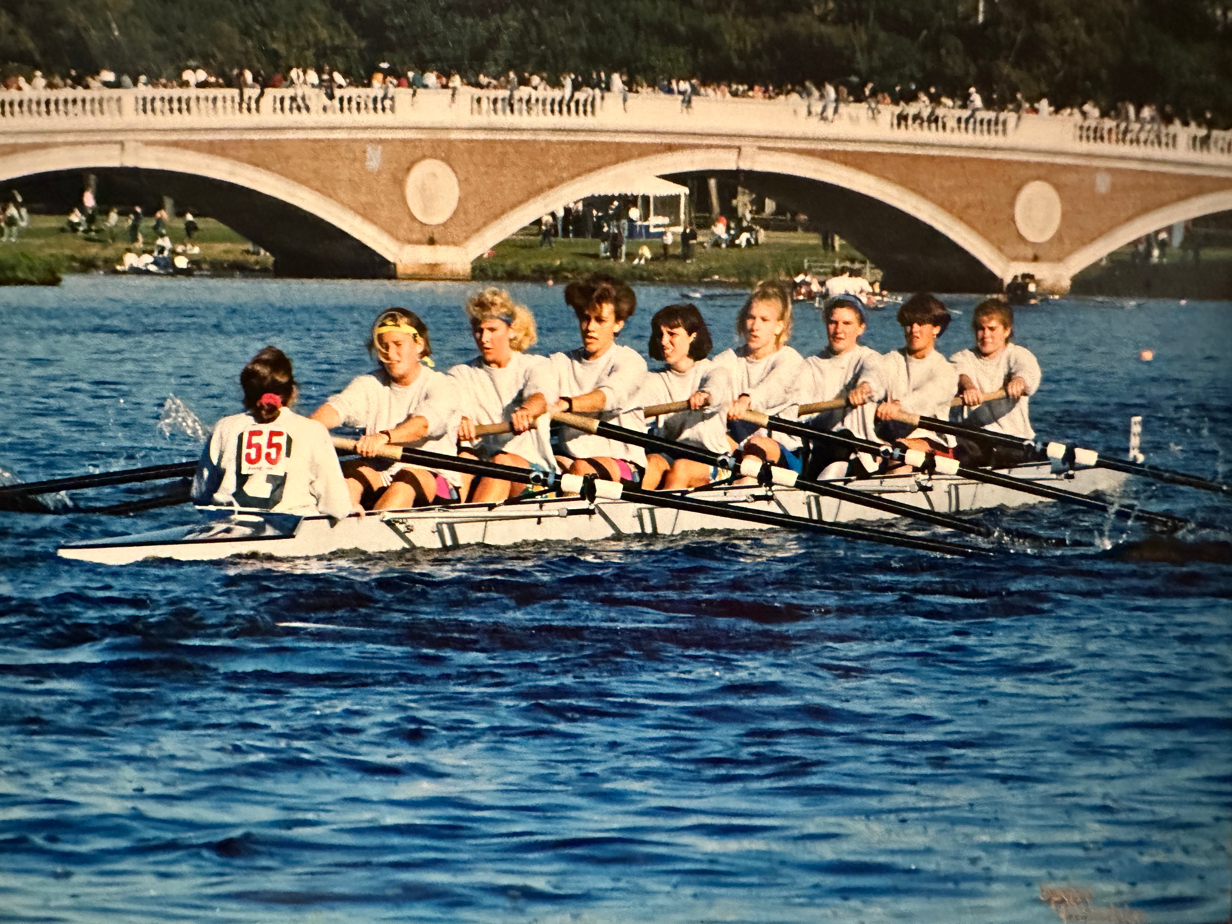 Nine women in 1990s clothing rowing a crew boat on a river with a low bridge in the background.