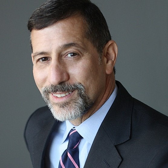 A man with brown hair and a brown and grey goatee wearing a dark grey suit and light blue shirt with necktie.