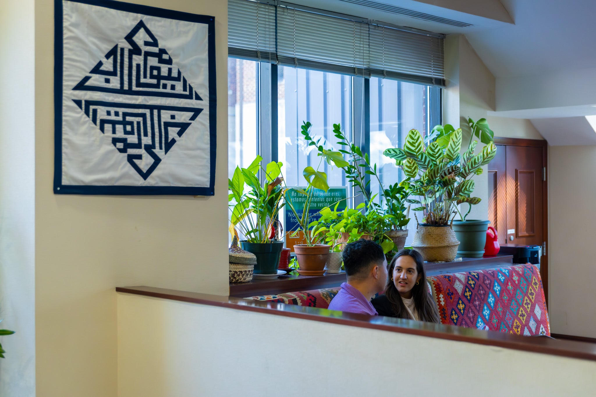 Two people sit in the CCAS lounge where the logo tapestry hangs in the forefront and plants sit in the background.