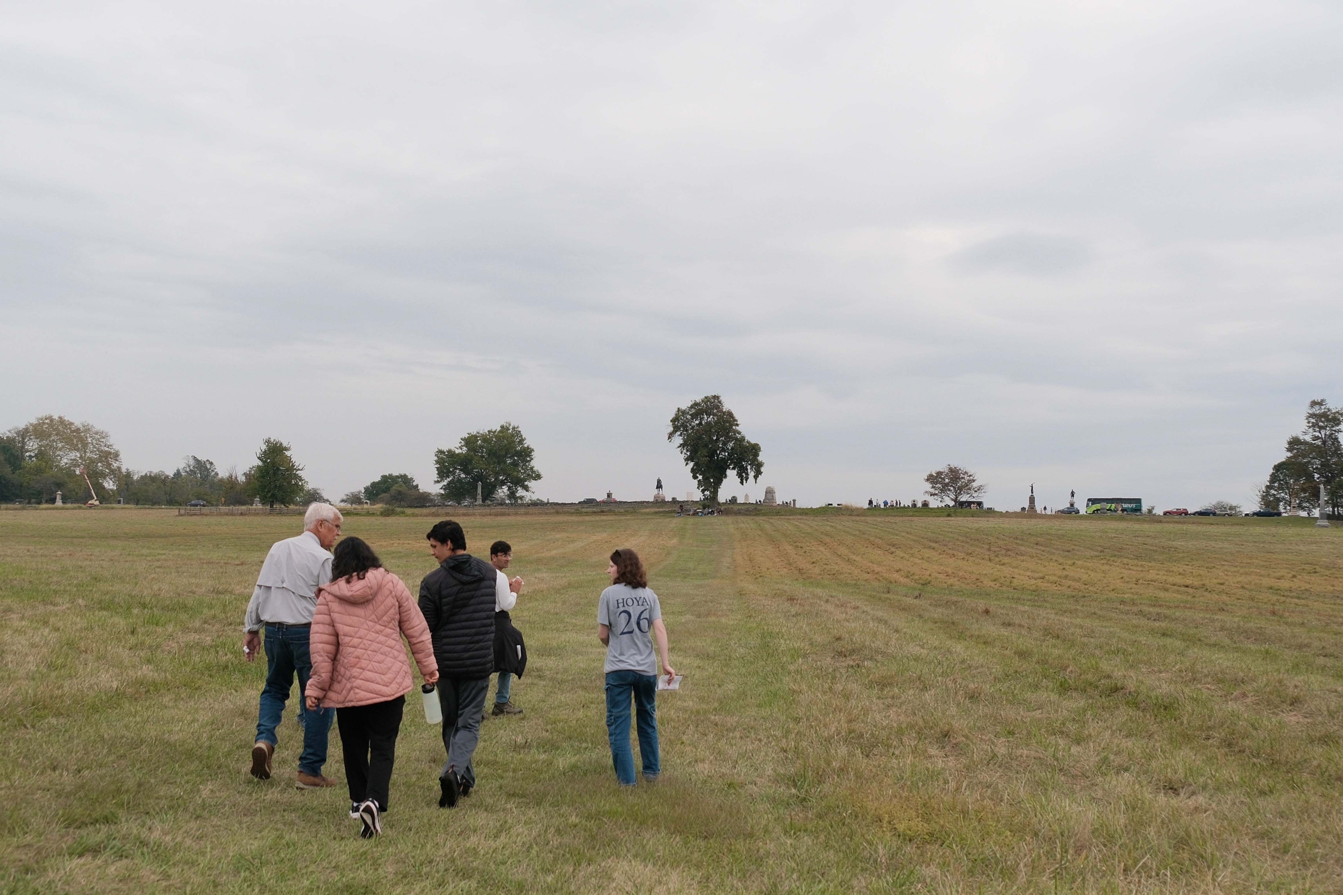 Four people walk across a field with their backs to the camera. The green field stretches to a series of monuments and trees at its edge.
