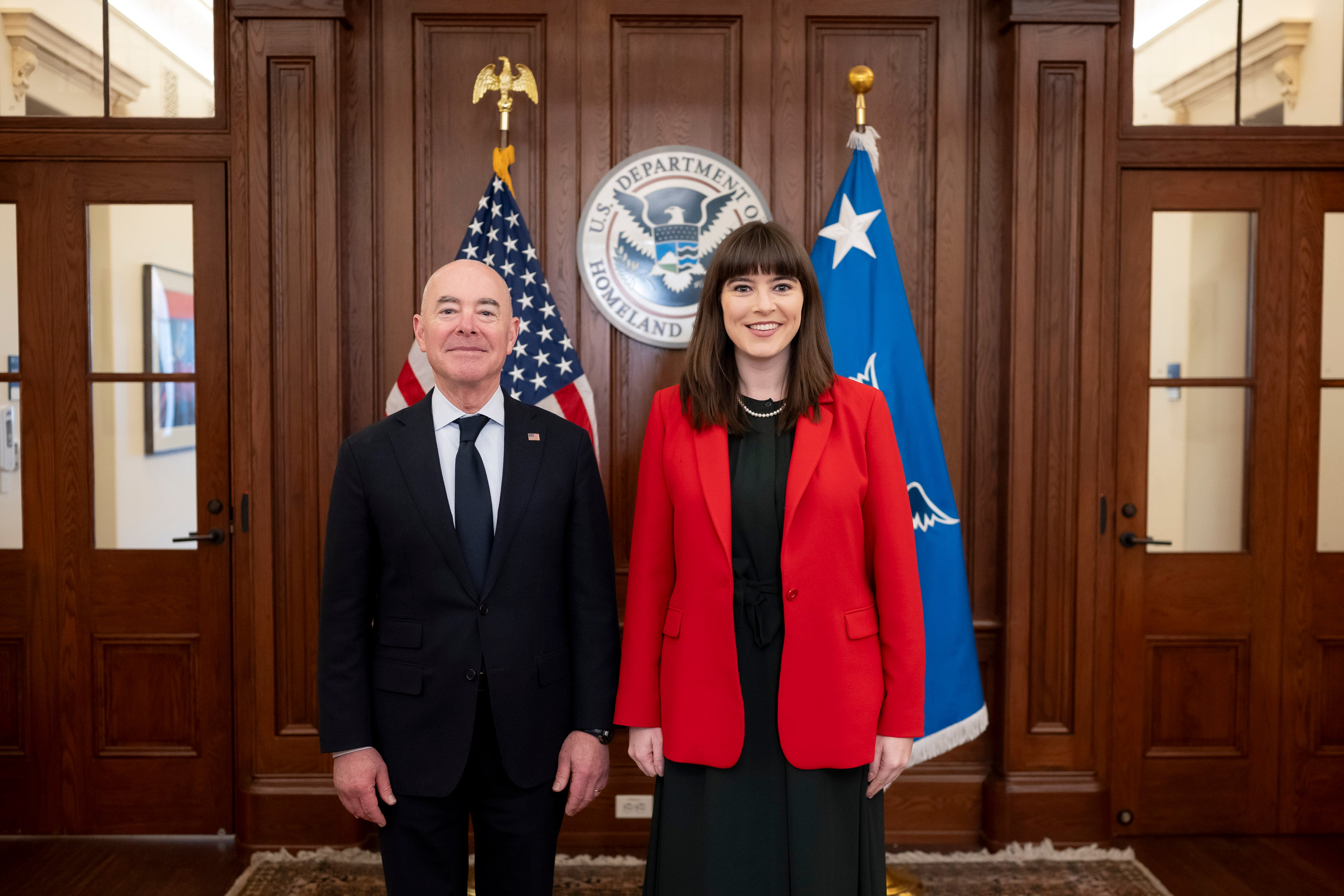 Two individuals standing and smiling in the Department of Homeland Security office, flanked by the U.S. and DHS flags.