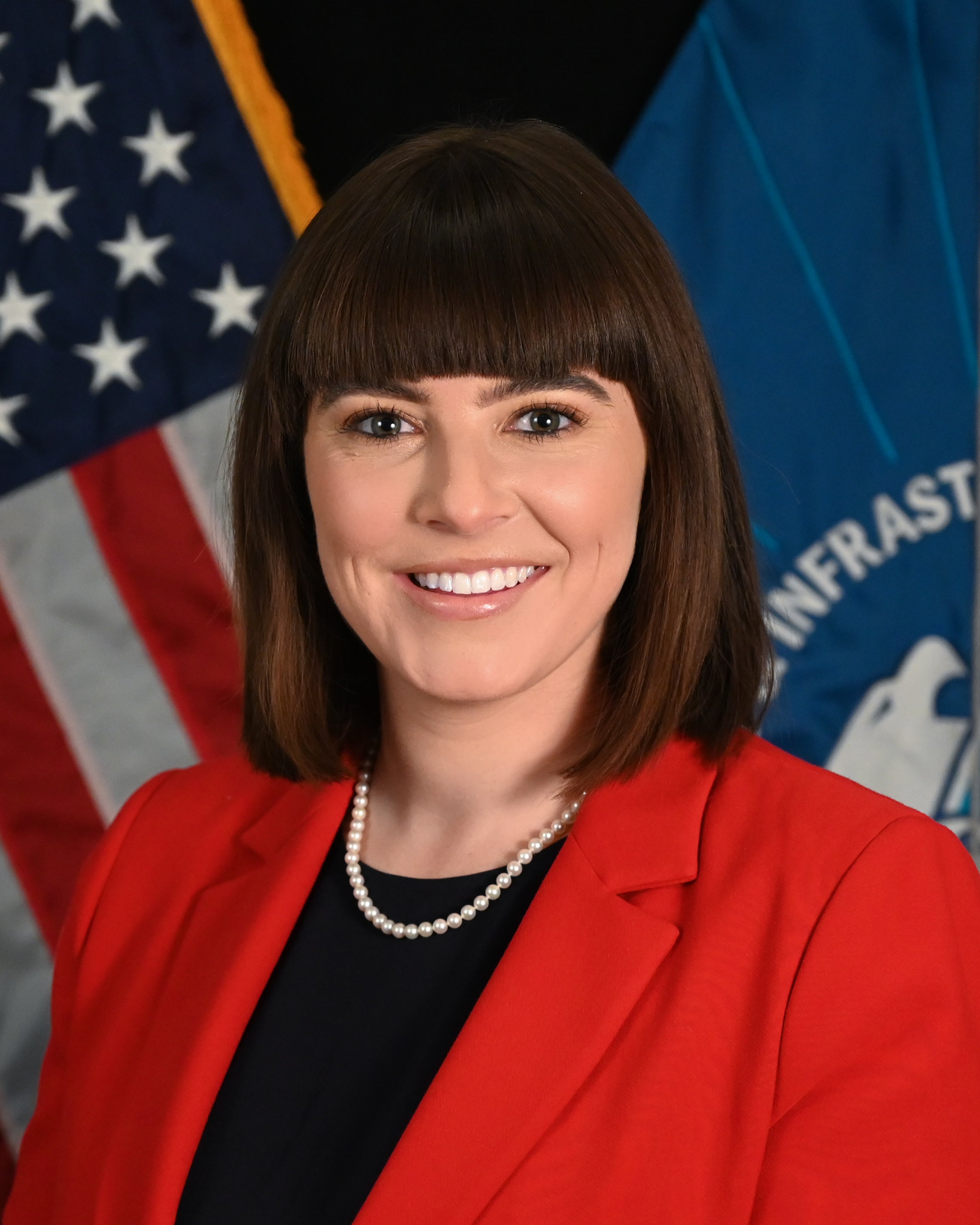 Official portrait of a person smiling, wearing a red jacket and a pearl necklace, with the United States flag and the Department of Homeland Security flag in the background.