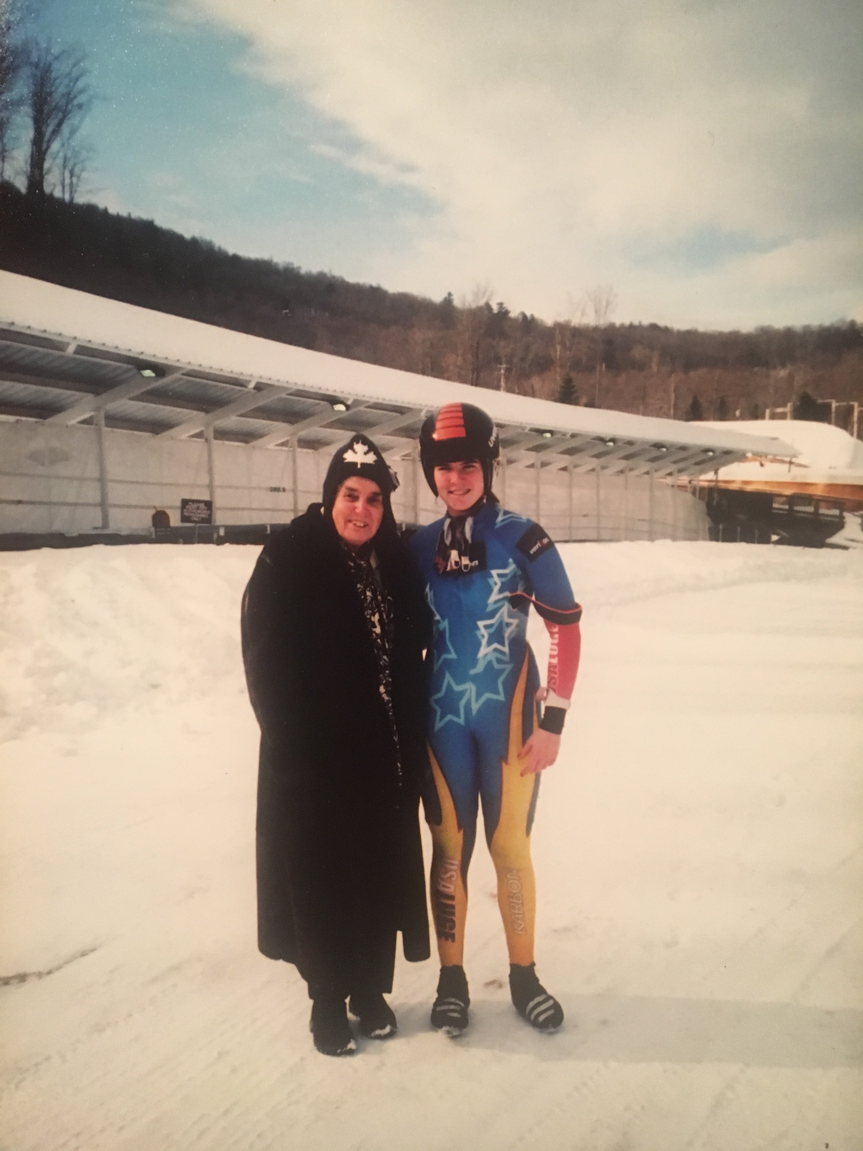 Two individuals in winter clothing standing on a snowy landscape, with one person wearing a colorful ski suit and the other in a dark coat and hat, near a fence with bare trees and a clear sky in the background.