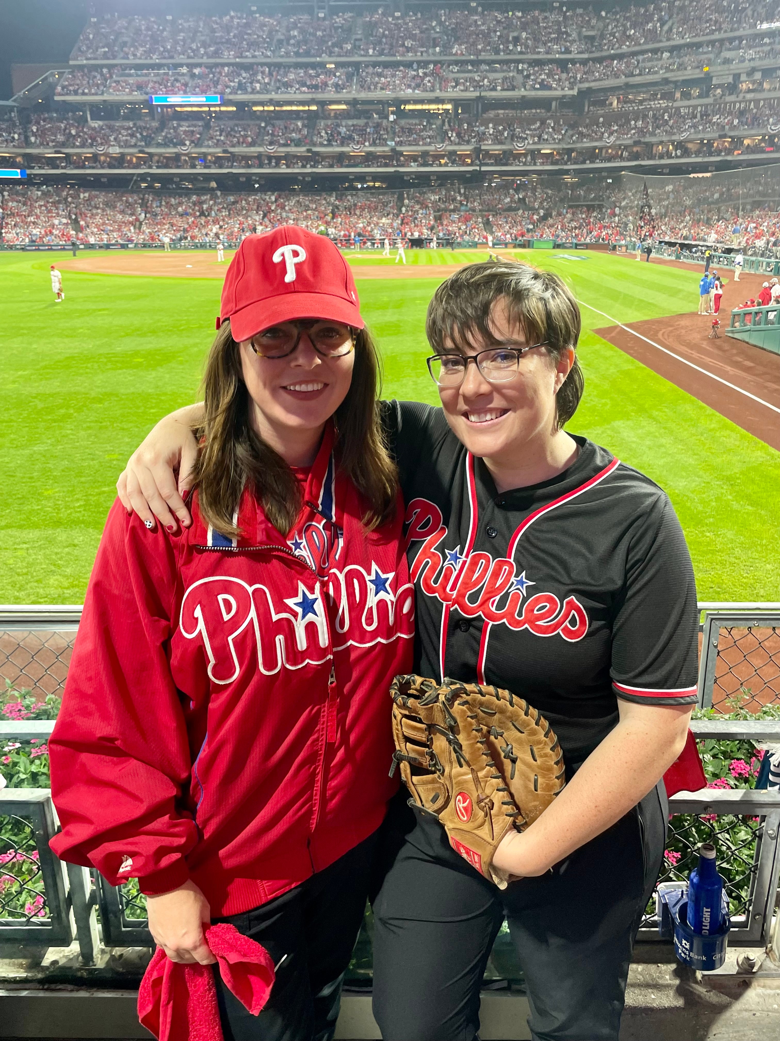 Two fans wearing Philadelphia Phillies attire smiling at a baseball game, with one holding a baseball glove, in a stadium filled with spectators.
