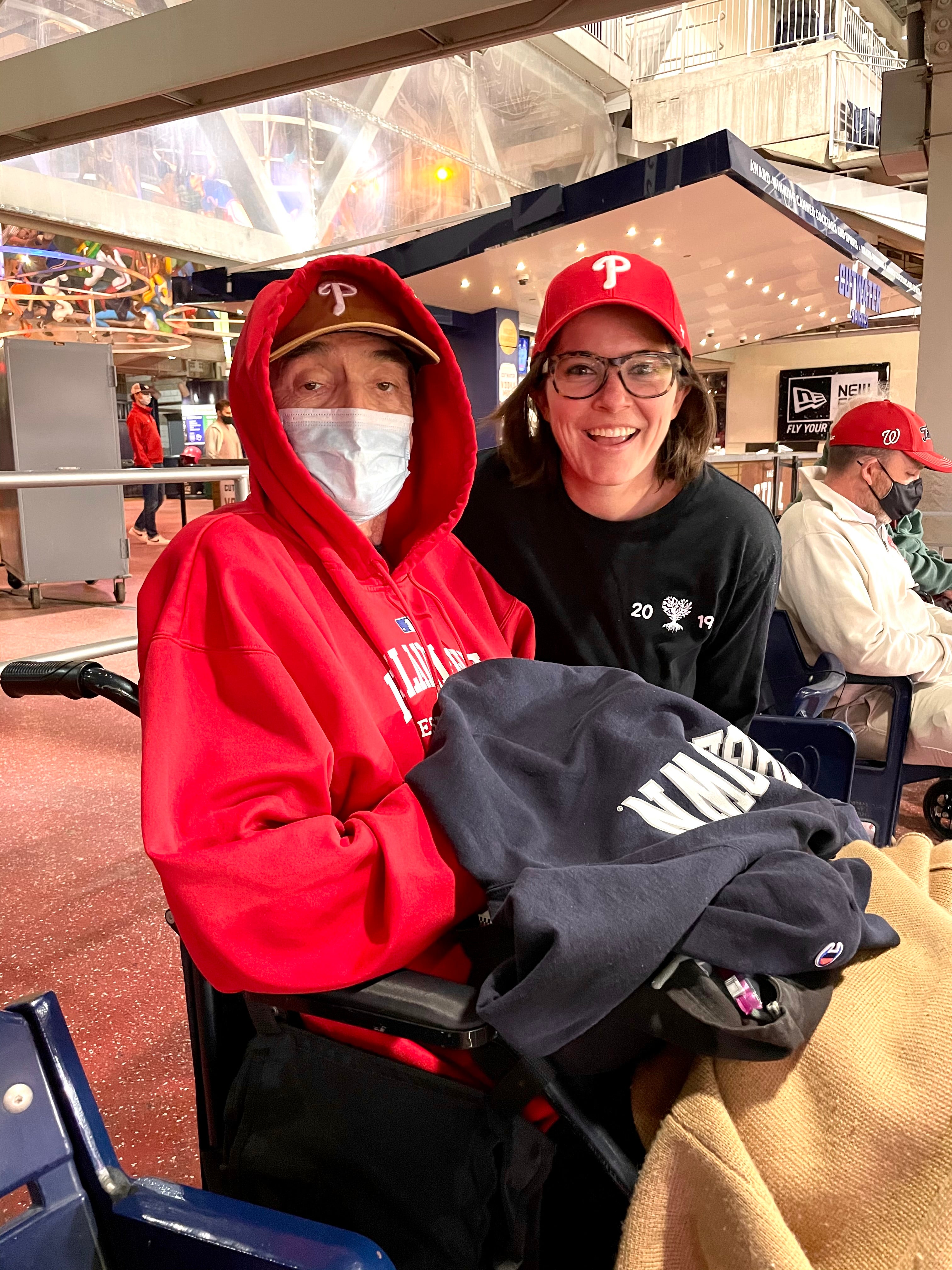 Two people wearing Philadelphia Phillies baseball caps, one in a red hoodie and mask seated in a wheelchair, and the other in a black t-shirt, smiling at a baseball stadium.