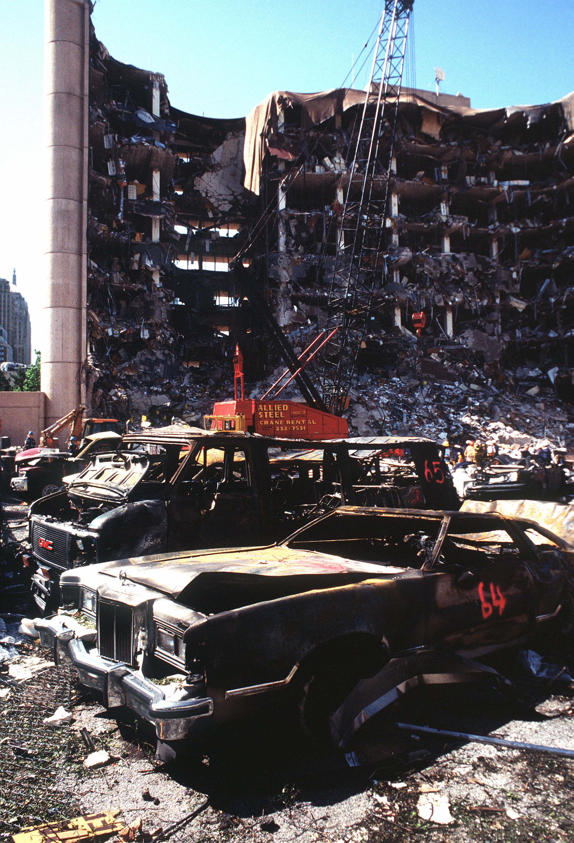 A multi-story, bombed out building with a crane in front of it. Foreground shows a burned out sedan and a burned out van.