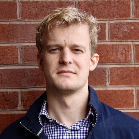 A blond man wearing a blue checked shirt and a blue windbreaker and standing in front of a brick wall.