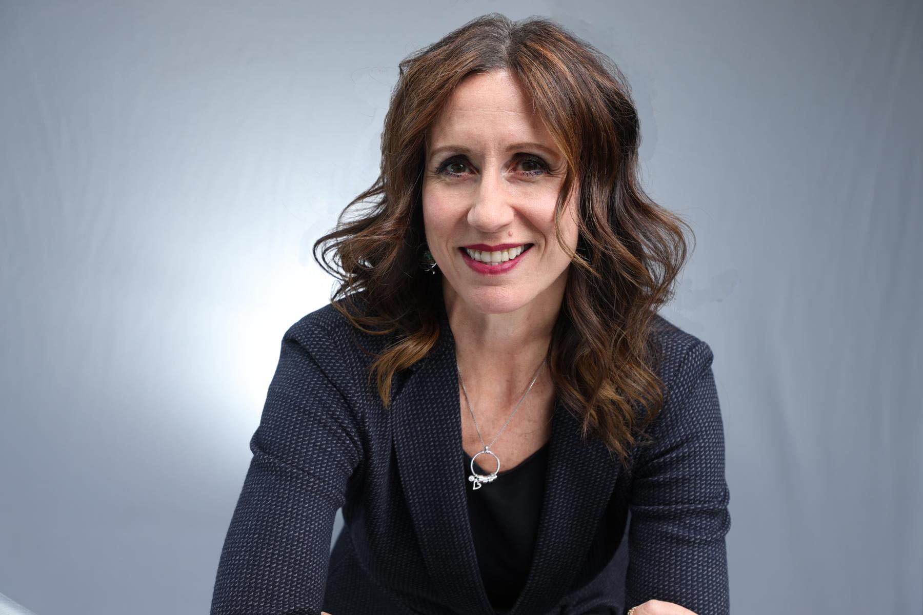 A woman with red-brown hair wearing a dark grey suit with a black shirt and silver necklace.