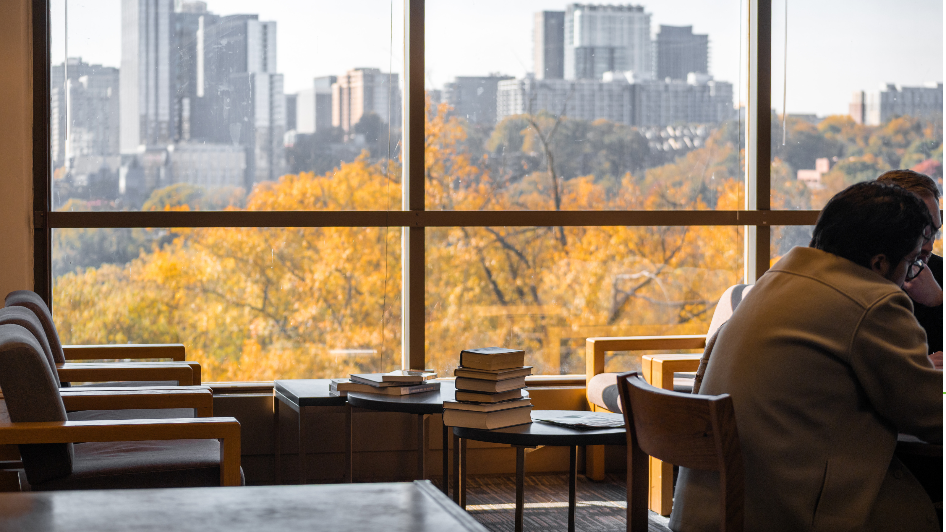 Person studying with books at a table by a large window overlooking a cityscape with autumn trees.