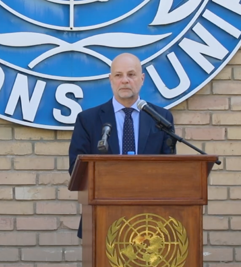 A person stands at a podium with microphones, speaking at an event in front of a United Nations logo.