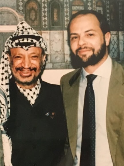 Two individuals smiling, standing side by side against a backdrop of decorative tiles. On the left is Yasser Arafat wearing a traditional keffiyeh.