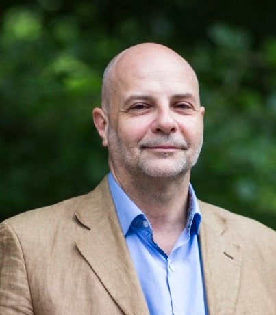 Portrait of an individual with a shaved head, wearing a beige blazer and blue shirt, smiling softly against a blurred green foliage background.