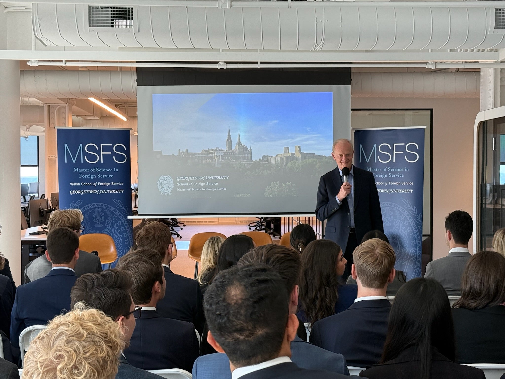 Man holding a microphone presenting to a room full of people. All wearing formal work attire. Behind him a slide show.