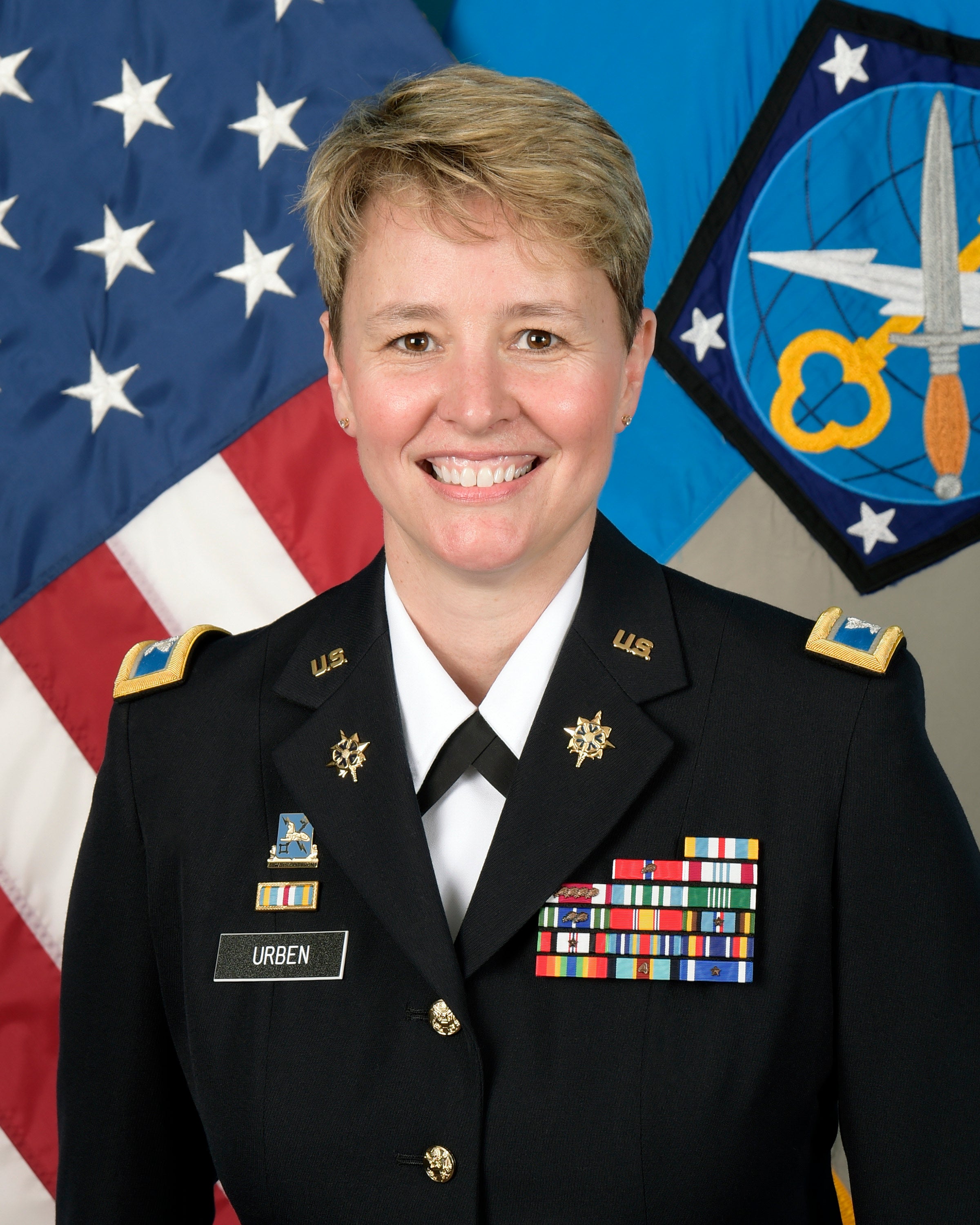 Portrait of a smiling U.S. military officer in uniform, adorned with medals, standing in front of the U.S. flag and a Space Force emblem.