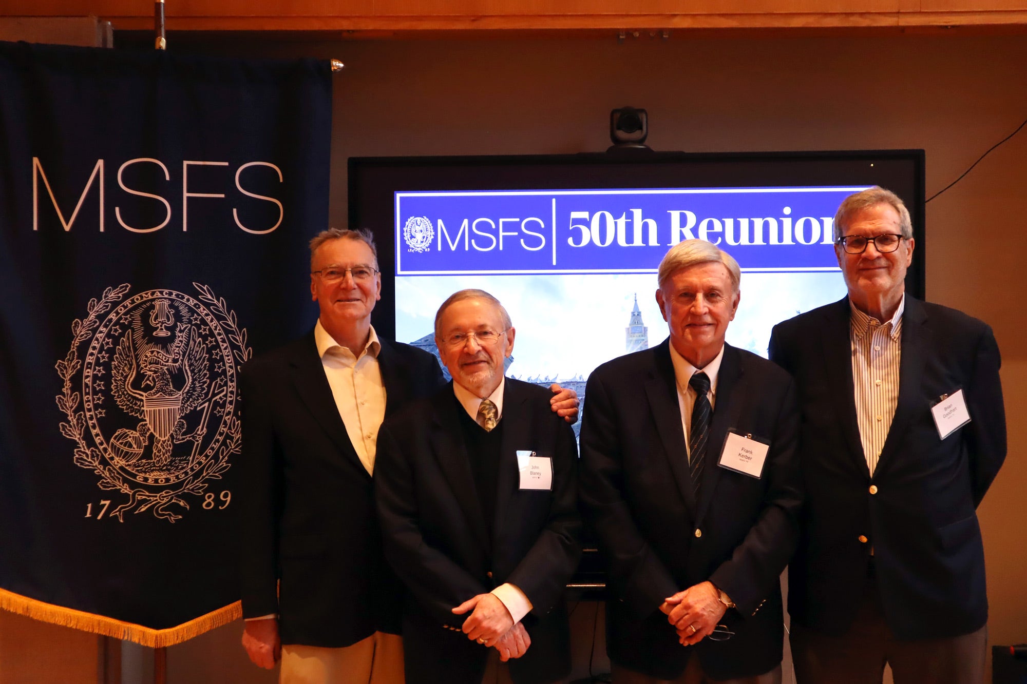 Four men with grey hair, wearing suits, standing together. Behind them a screen stating "MSFS 50th Reunion"
