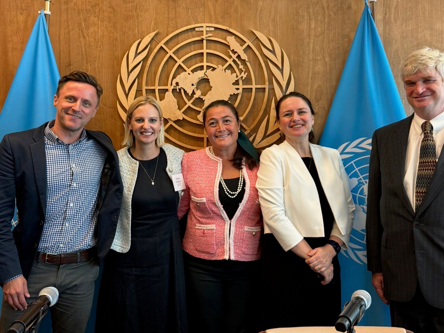 Five people standing in front of the United Nations logo, smiling, wearing business formal clothes.