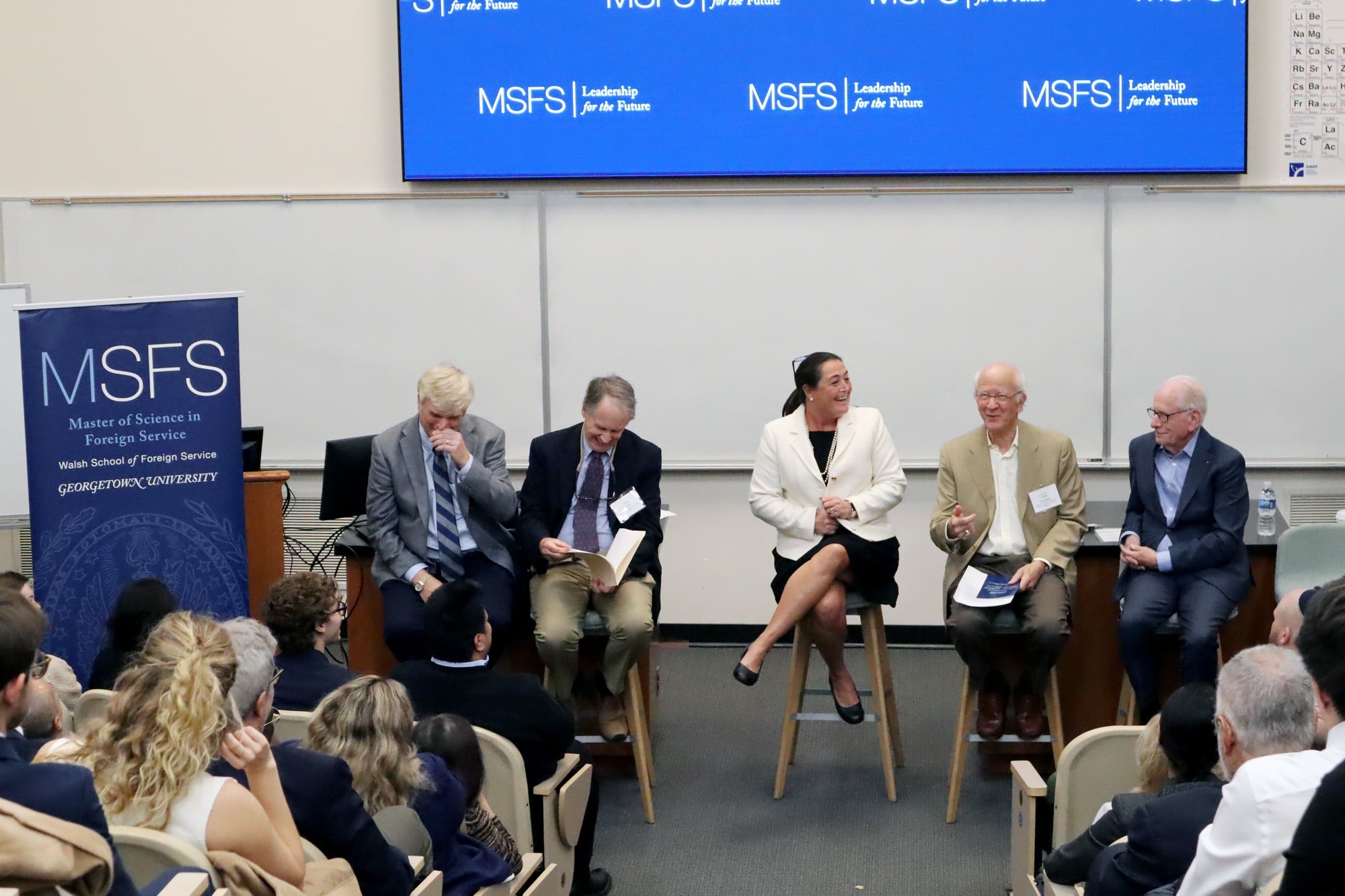 Five panelists sitting in high chairs, speaking in front of full a classroom. Next to them a banner that says MSFS Master of Science in Foreign Service.